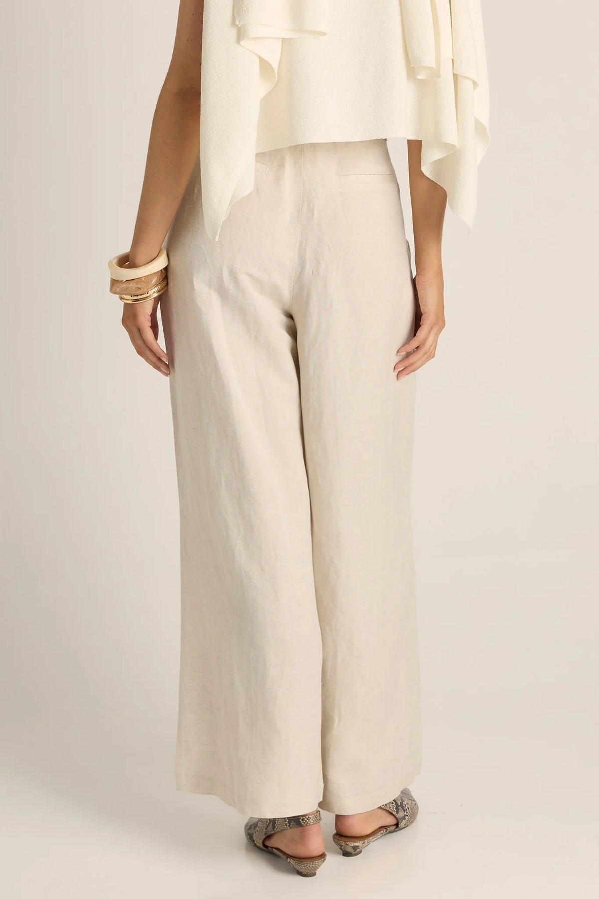 Woman shown from the back wearing Fate & Becker high-waisted, wide-leg trousers in a light cream or off-white linen-like fabric, paired with a matching draped top and flat sandals, against a plain light beige studio background.