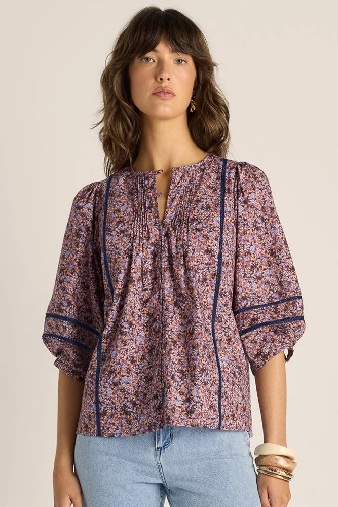 Image of A woman wearing a Fate & Becker short-sleeve floral blouse with dark blue piping and a split neckline, paired with light blue jeans, against a plain beige background.