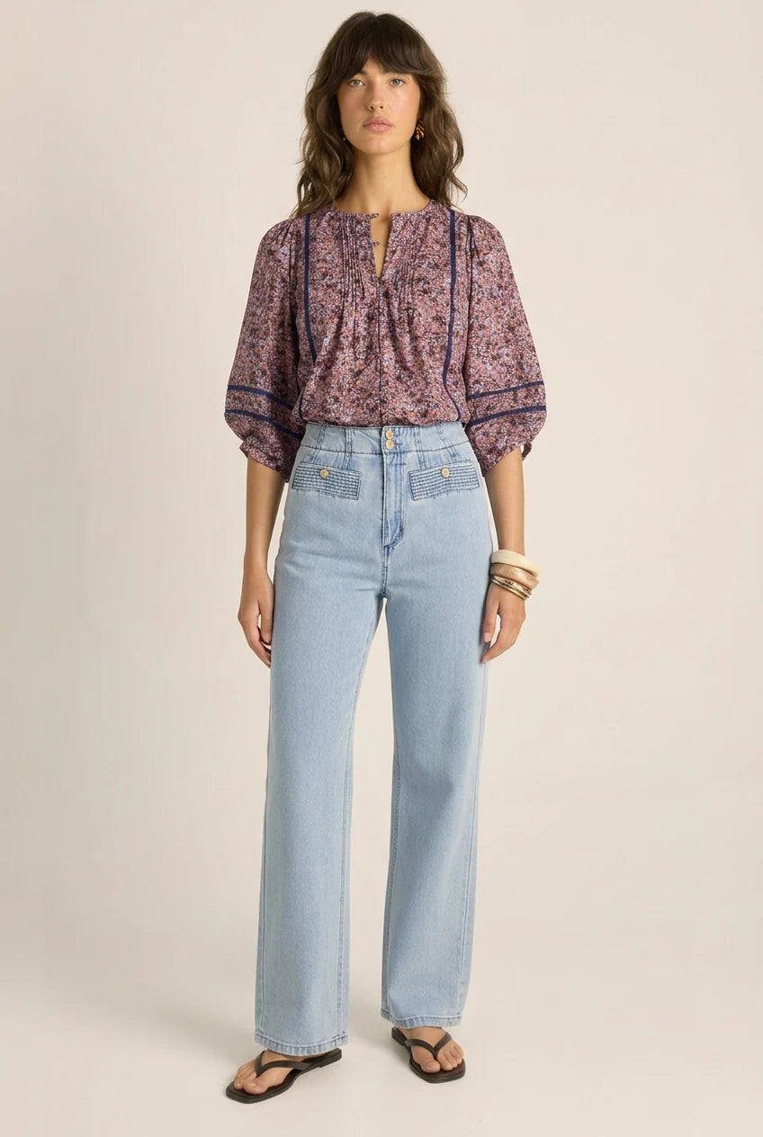 Image of A woman modeling a Fate & Becker purple and pink floral patterned blouse with puff sleeves and light-wash blue wide-leg jeans, standing against a plain beige studio background.