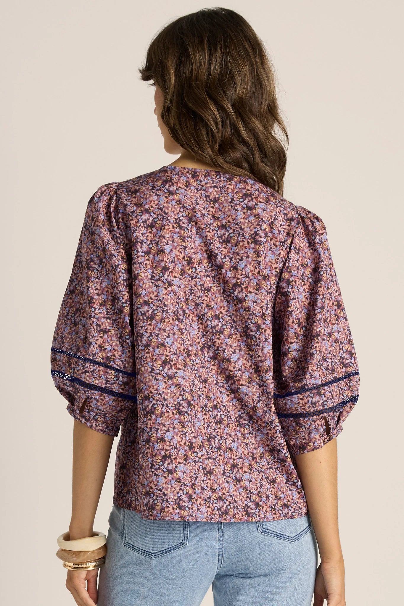 Image of A woman shown from the back wearing a Fate & Becker short-sleeve blouse with a dense floral print in dusty pink, purple, and orange tones, paired with light blue jeans against a solid light beige background.