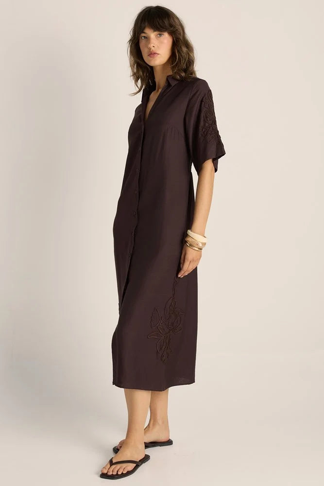 Image of A woman modeling a Fate & Becker dark brown, short-sleeved button-up shirt dress with embroidered details on the sleeves and lower skirt, paired with black flip-flops against a light beige studio background.