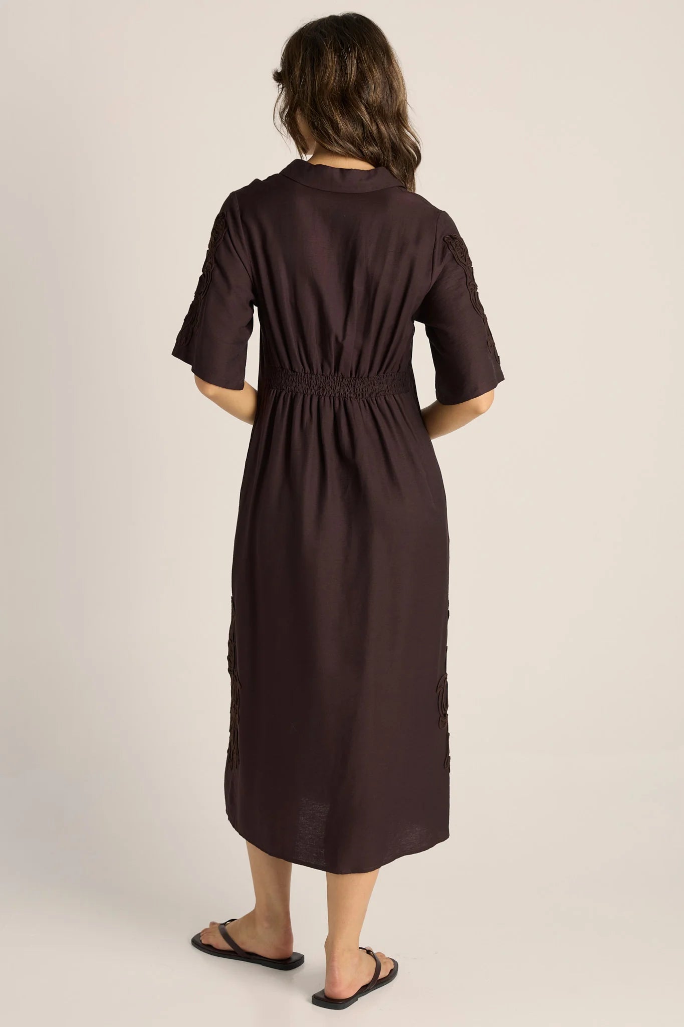 Image of A woman viewed from behind wearing a Fate & Becker dark brown short-sleeve midi dress with a gathered waist and side slit details, paired with black flip-flop sandals, against a plain light beige studio background.