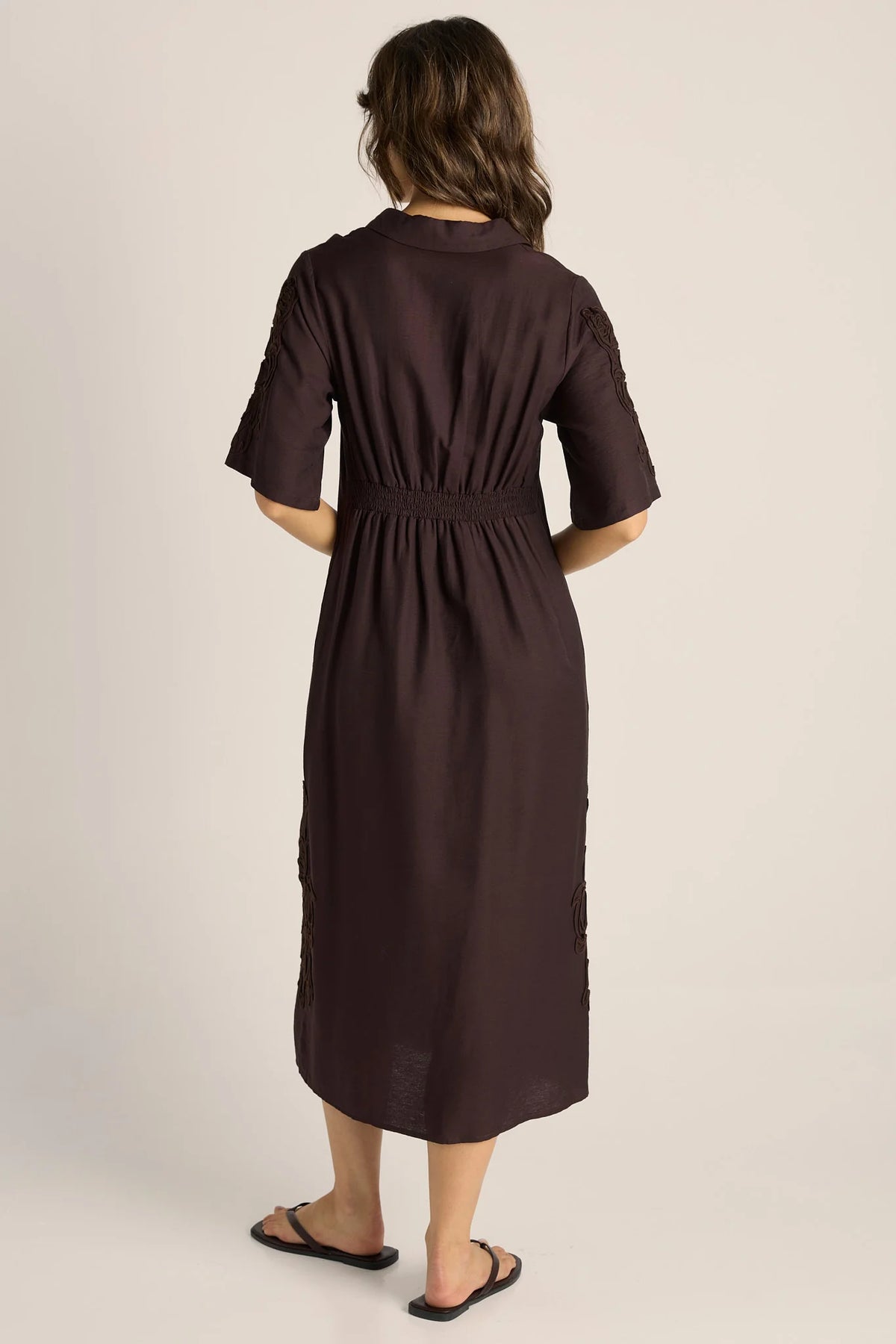 A woman viewed from behind wearing a Fate & Becker dark brown short-sleeve midi dress with a gathered waist and side slit details, paired with black flip-flop sandals, against a plain light beige studio background.