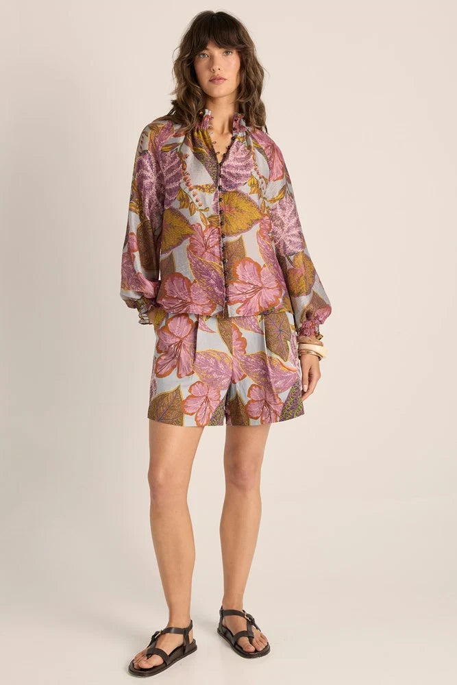 Image of A model stands against a plain beige studio background wearing a Fate & Becker matching long-sleeve button-front top and shorts set with a pink, purple, and gold botanical print, paired with black strappy sandals.