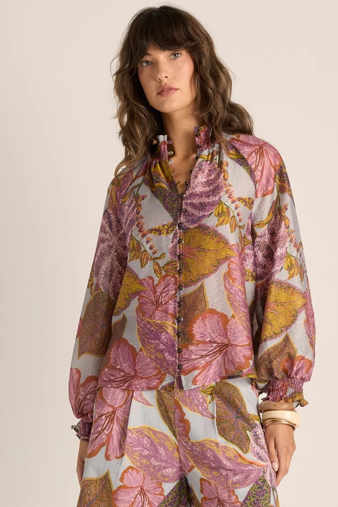 A woman wearing a Fate & Becker long-sleeve button-front blouse with a large botanical floral print in pink, purple, mustard yellow, and olive tones on a light base, photographed against a plain warm beige studio background.