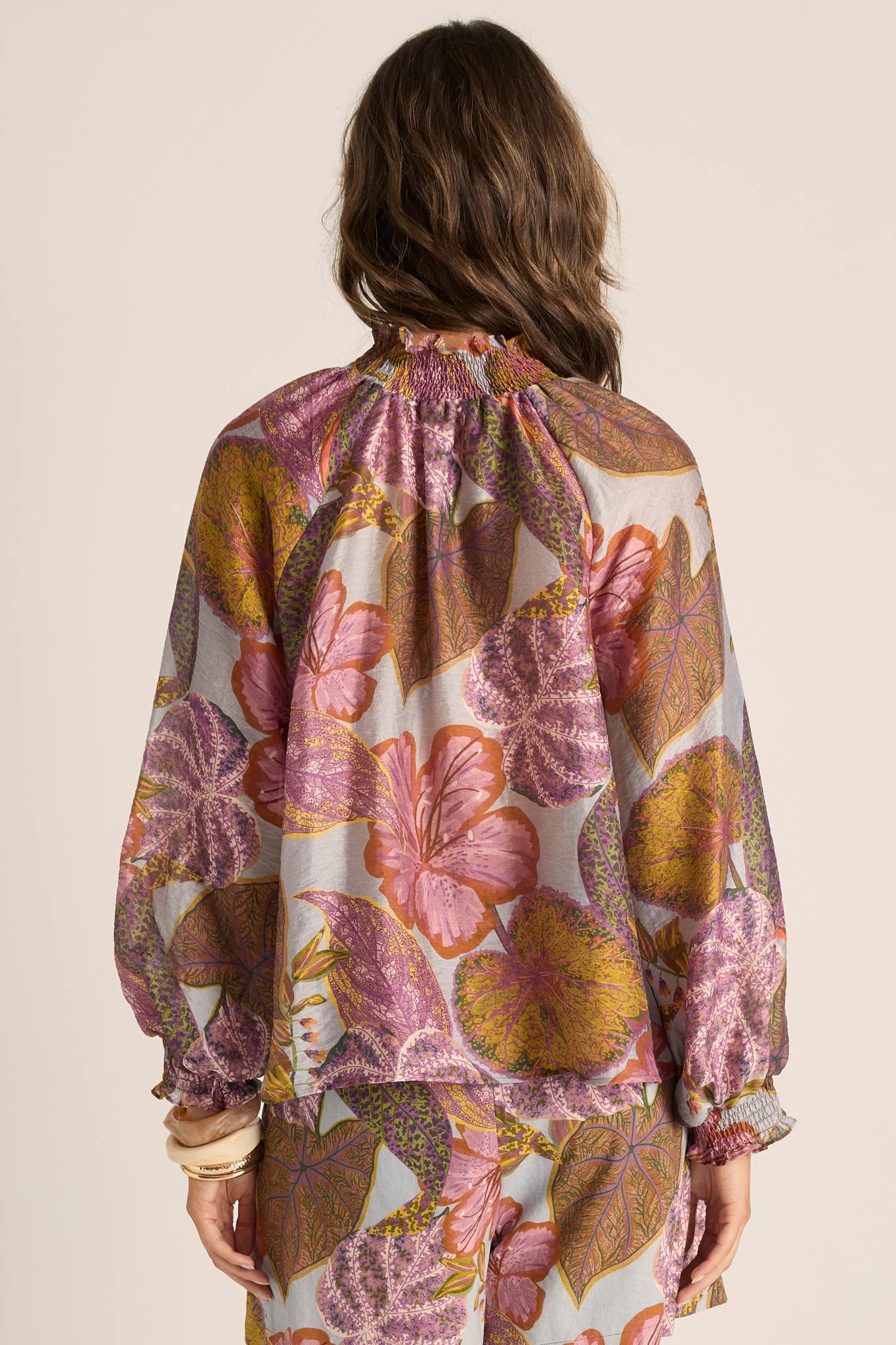Image of A woman shown from the back wearing a Fate & Becker loose, long-sleeve blouse with a large-scale floral and botanical print in pink, purple, mustard yellow, and brown on a light cream background.