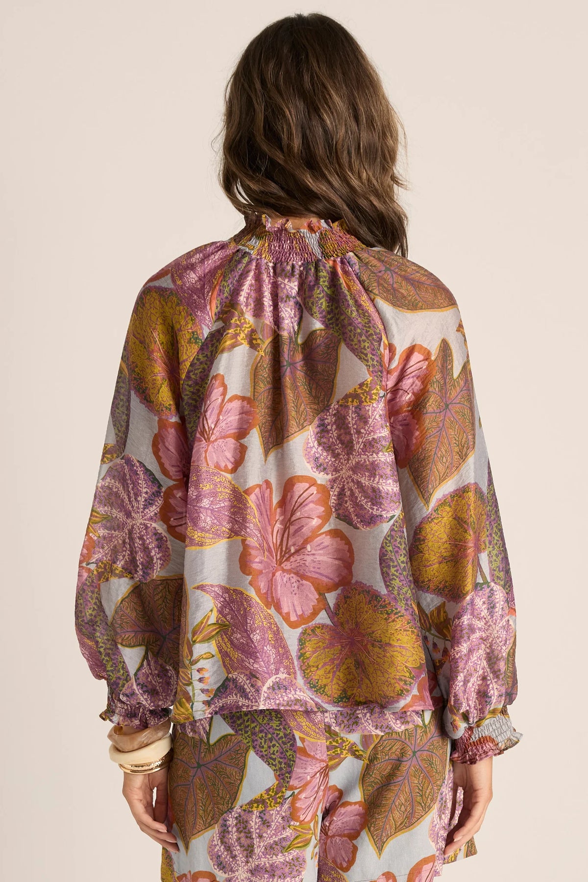 A woman shown from the back wearing a Fate & Becker loose, long-sleeve blouse with a large-scale floral and botanical print in pink, purple, mustard yellow, and brown on a light cream background.