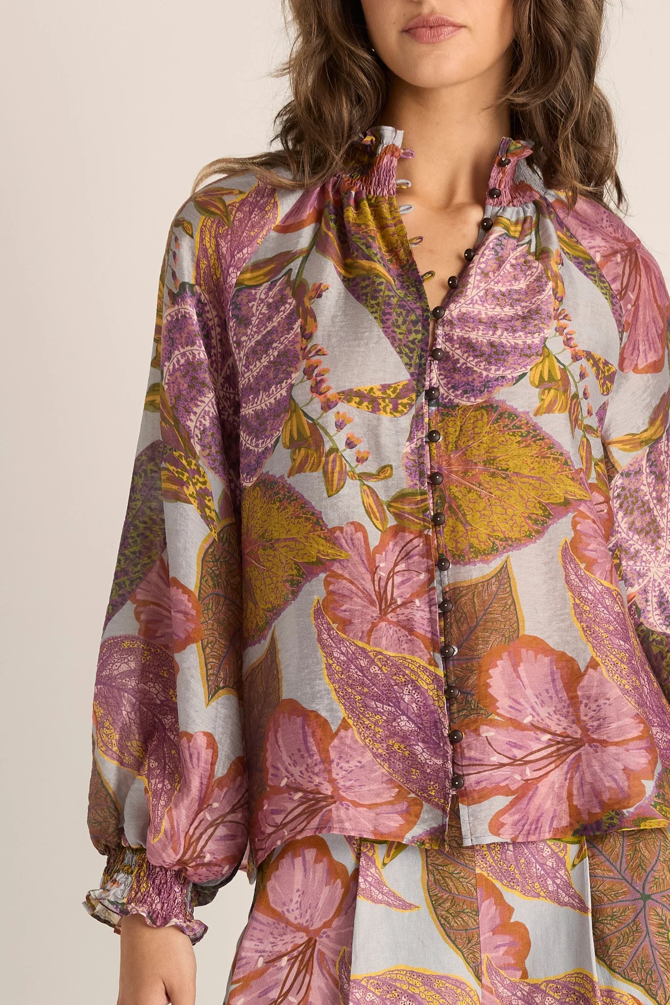 Image of A close-up of a woman wearing a Fate & Becker long-sleeve button-front blouse with a large floral print in pink, purple, mustard yellow, and olive tones on a light background.
