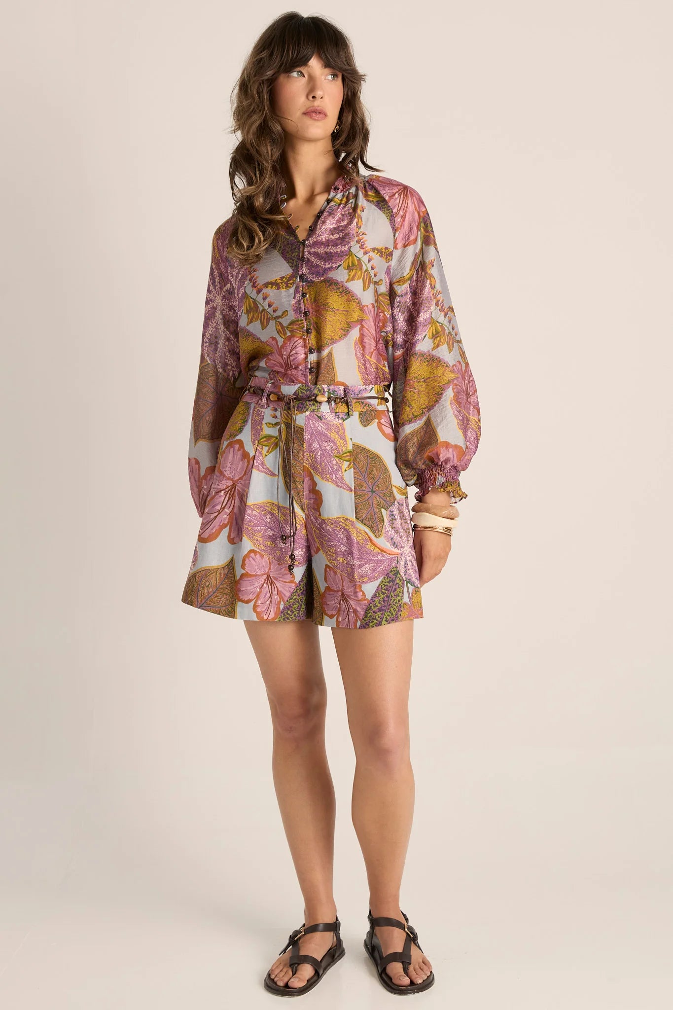 Image of A woman modeling a Fate & Becker long-sleeve floral shirt with a tie waist, shown against a plain beige studio background.