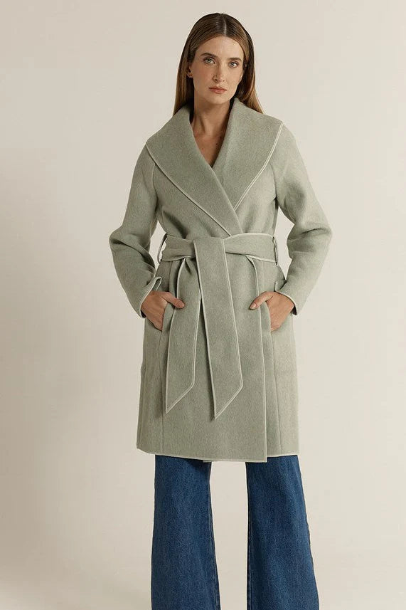 Woman wearing a Fate & Becker light green coat with a belt, standing against a plain background