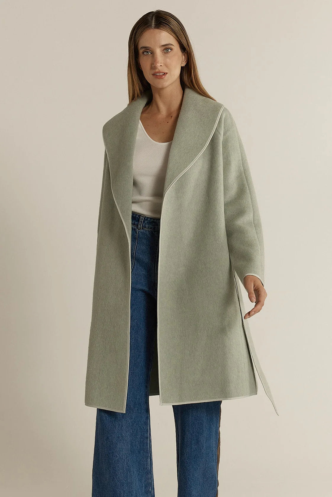 Image of Woman wearing a Fate & Becker light green coat over a white top and blue jeans on a beige background