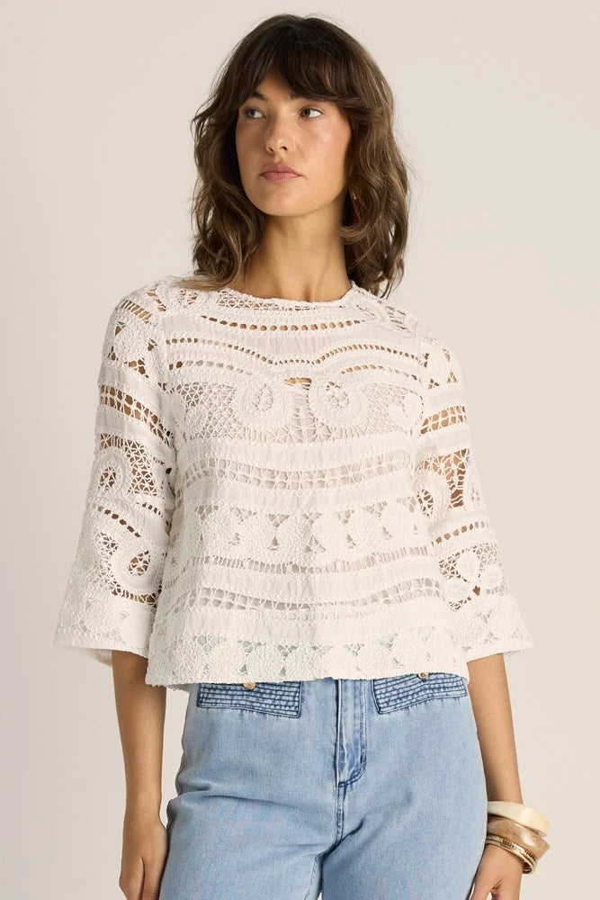 Image of A woman wearing a Fate & Becker white crochet knit top with three-quarter sleeves and light blue jeans, standing against a plain beige studio background.