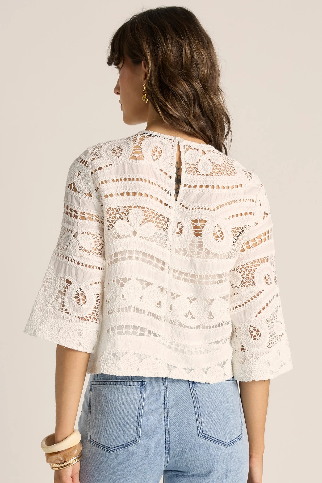 Image of A woman shown from the back wearing a Betty Basics white lace three-quarter sleeve crop top and light blue jeans against a plain beige studio background.