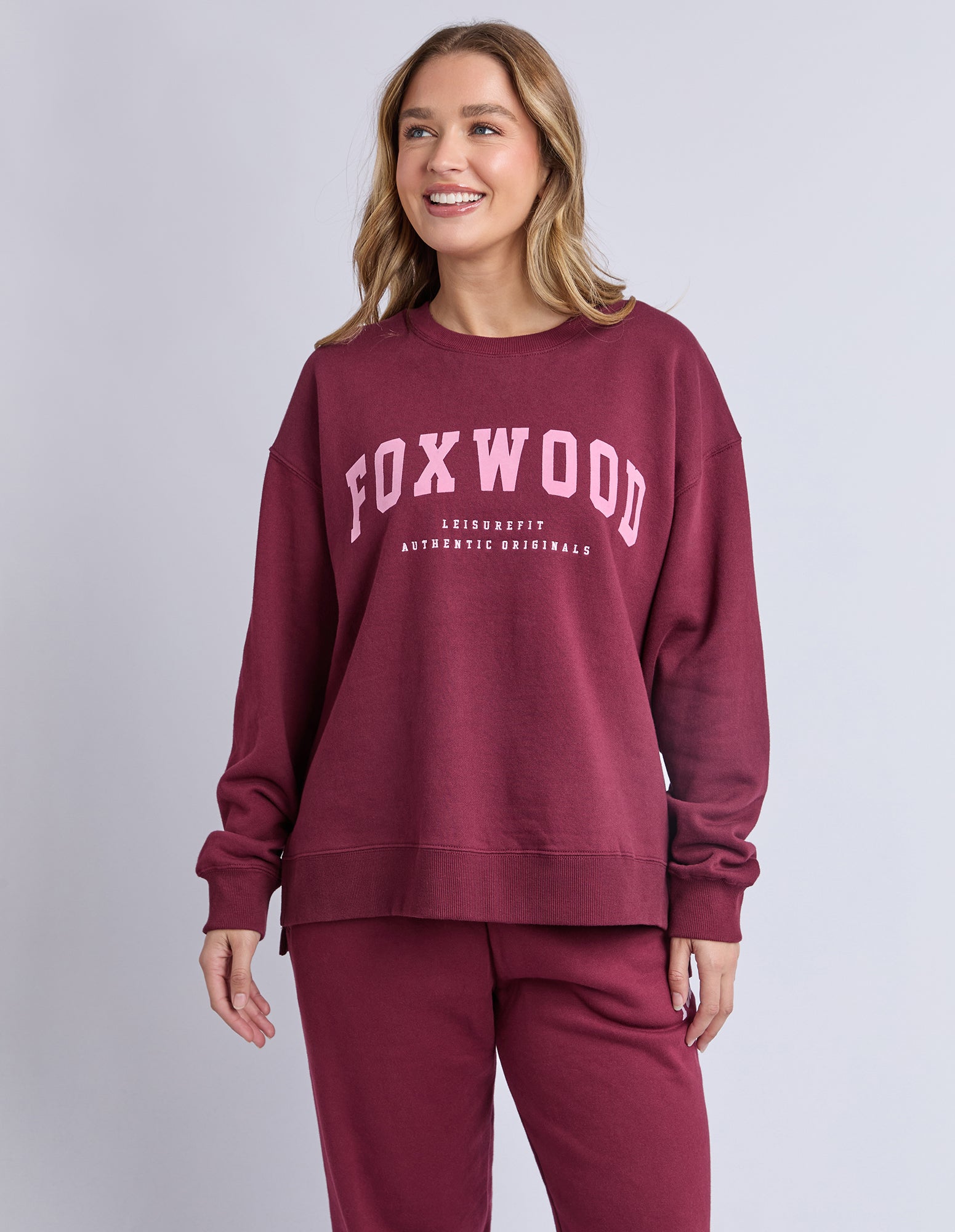 Image of Woman wearing a maroon jumper with 'FOXWOOD' printed on it in pink against a light grey background
