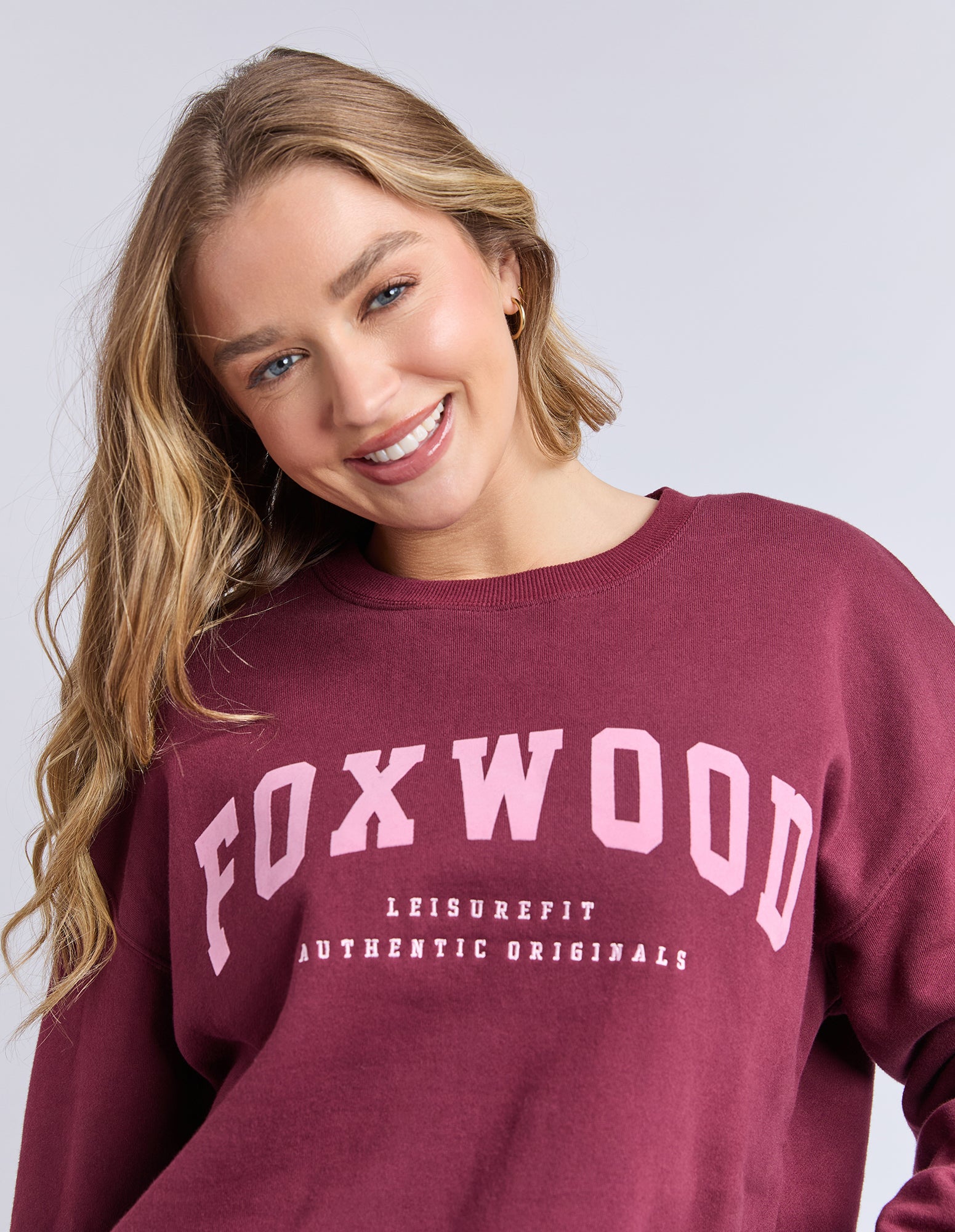 Image of Blonde woman wearing a maroon jumper with 'FOXWOOD' branding in pink on a light grey background