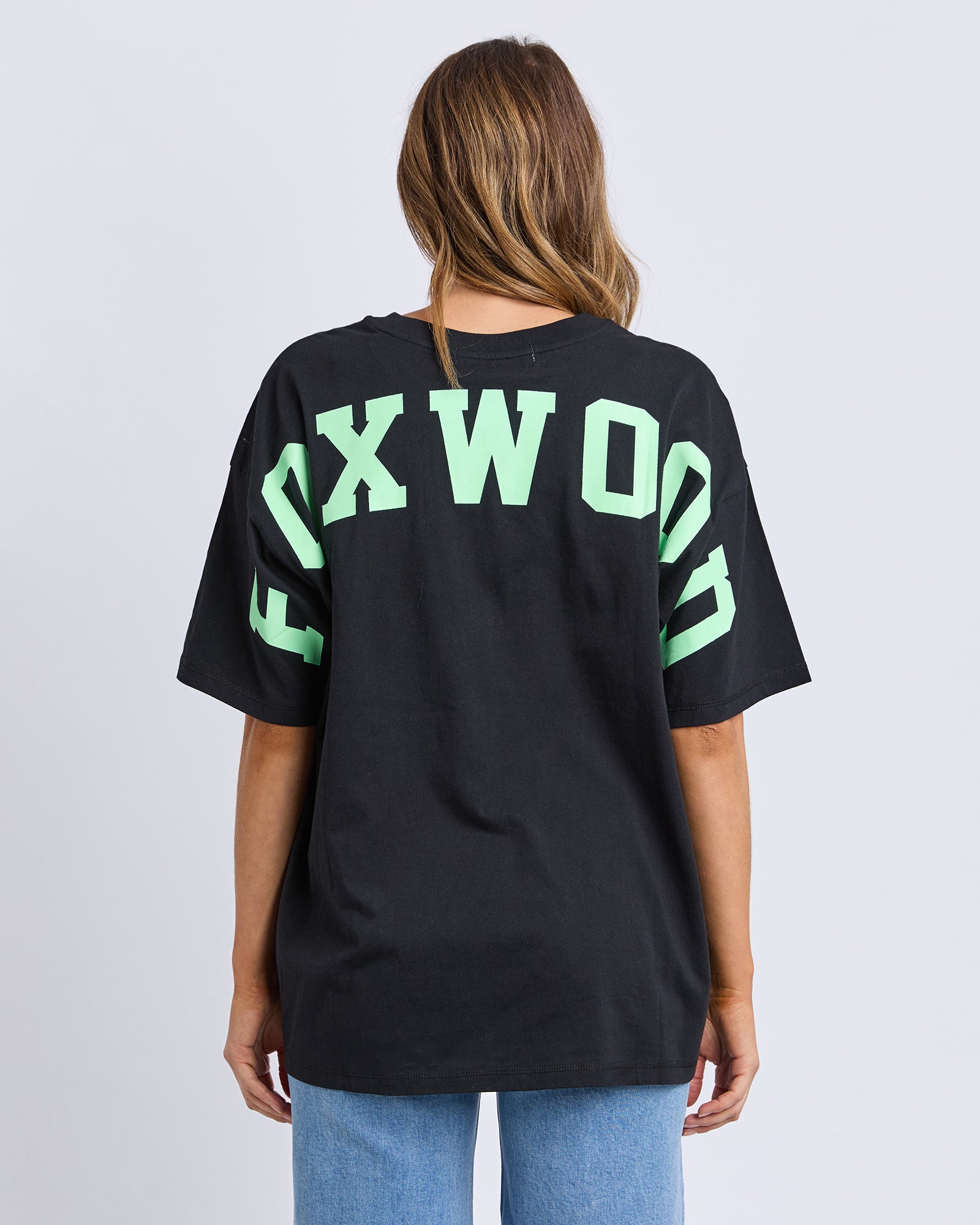 Collection of Woman wearing a Foxwood black t-shirt with 'FOXWOOD' printed on the back in green against a light grey background in a gallery layout