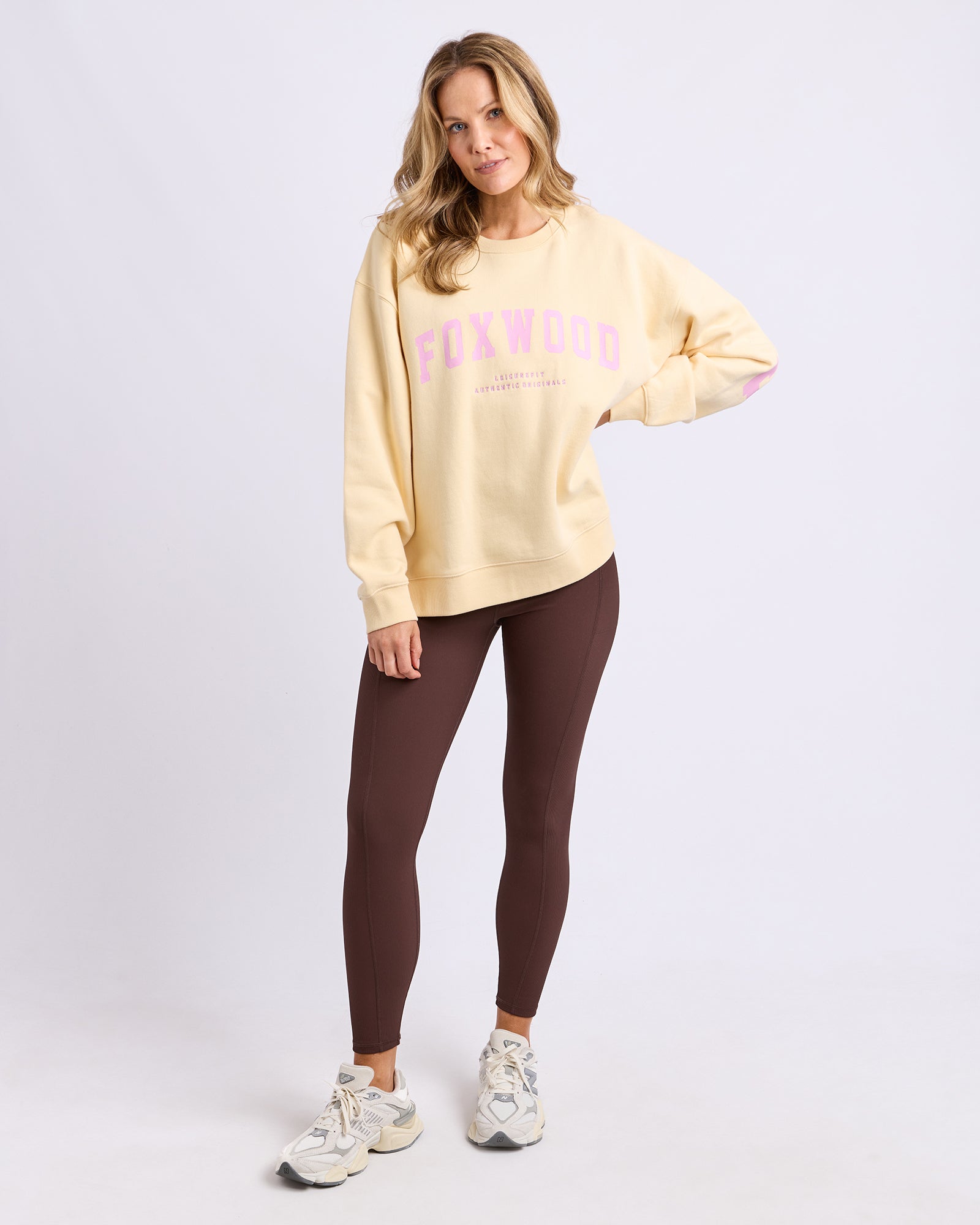 Image of Woman wearing a Foxwood light yellow jumper with pink text and brown leggings on a white background