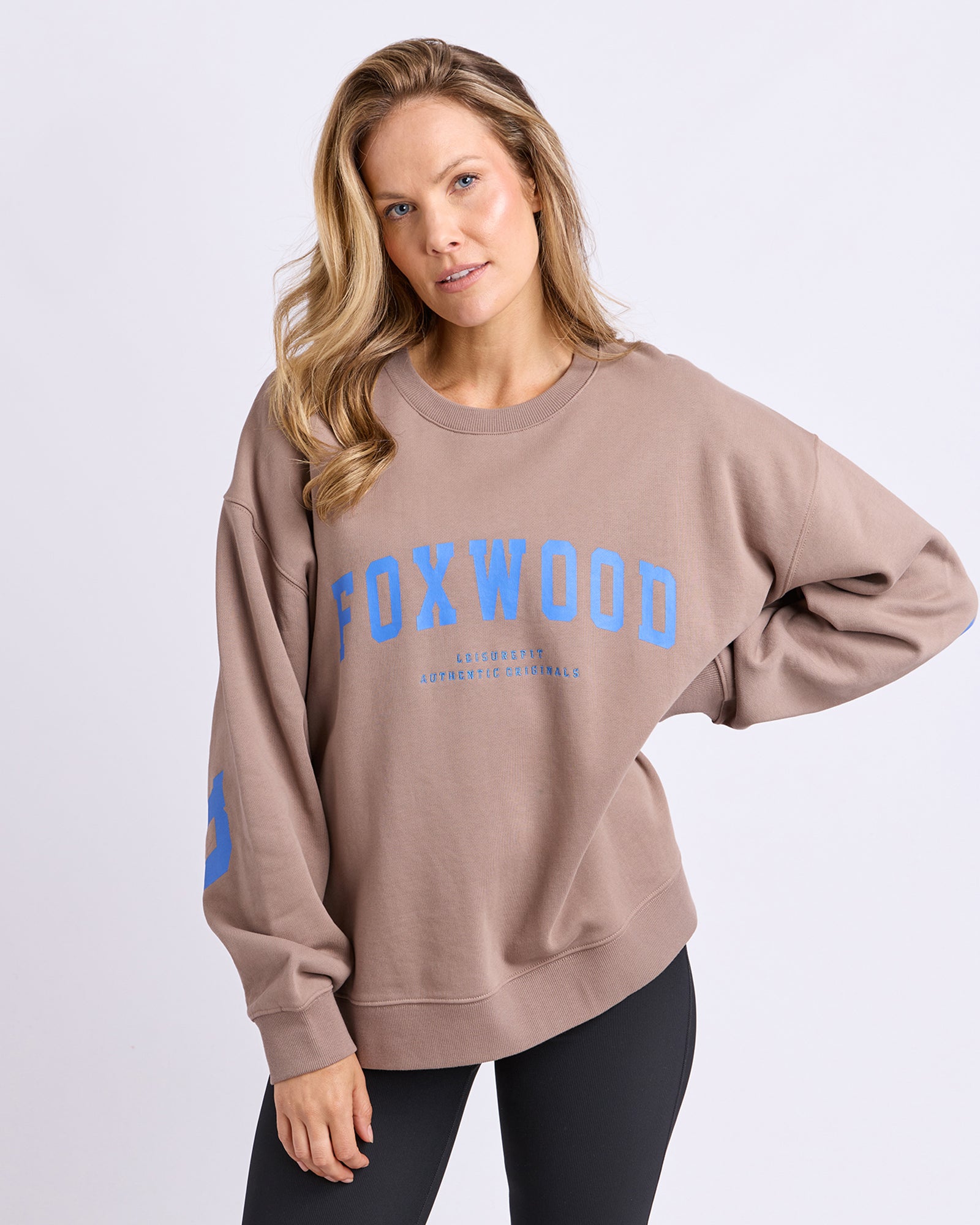 Woman wearing a brown crew jumper with 'FOXWOOD' printed in blue on a light gray background