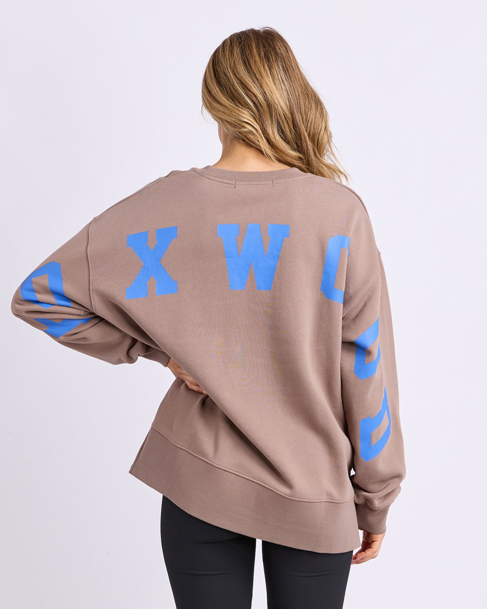 Back view of a woman wearing a Foxwood brown jumper with blue letters on a plain background