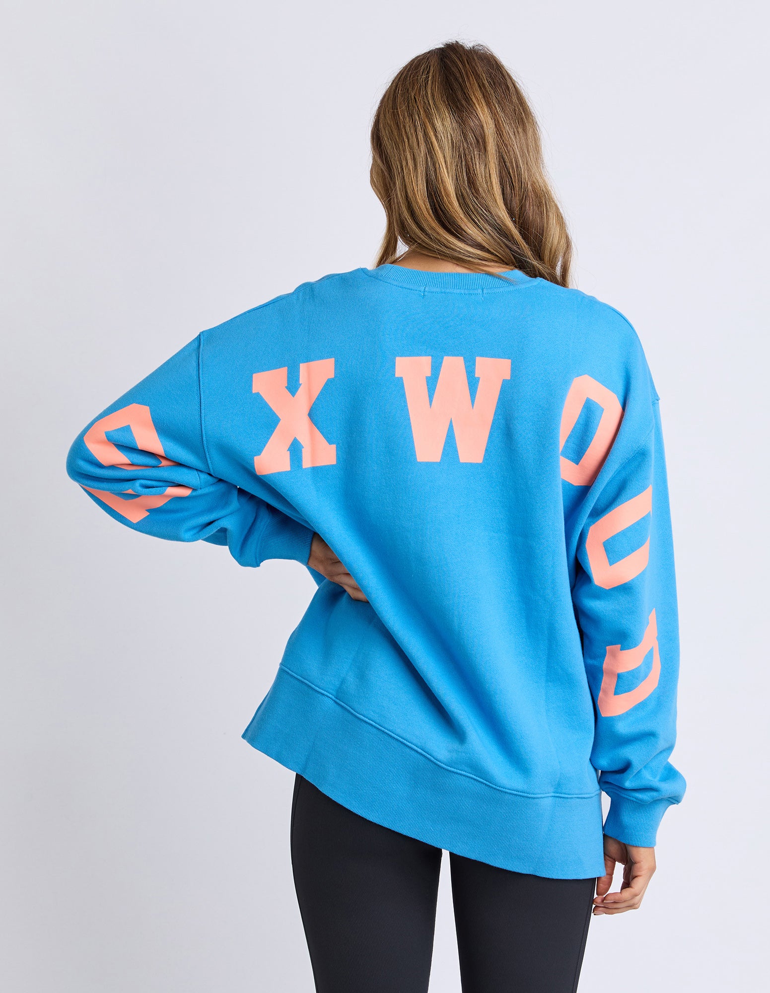 Back view of a woman wearing a Foxwood bright blue jumper with orange letters on a plain background