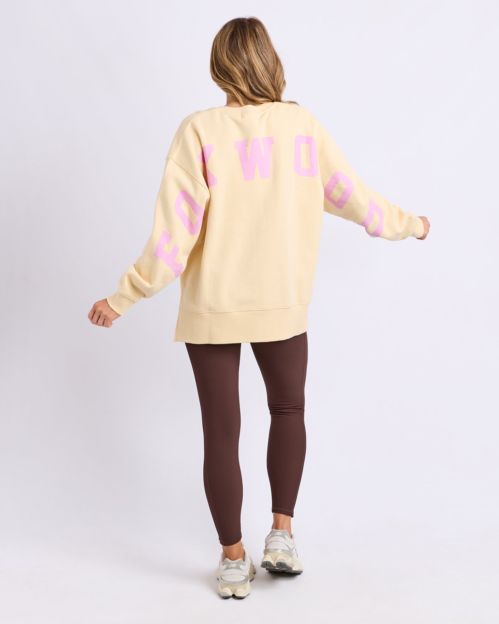 Back view of a woman wearing a Foxwood light yellow jumper with pink text and brown leggings on a white background