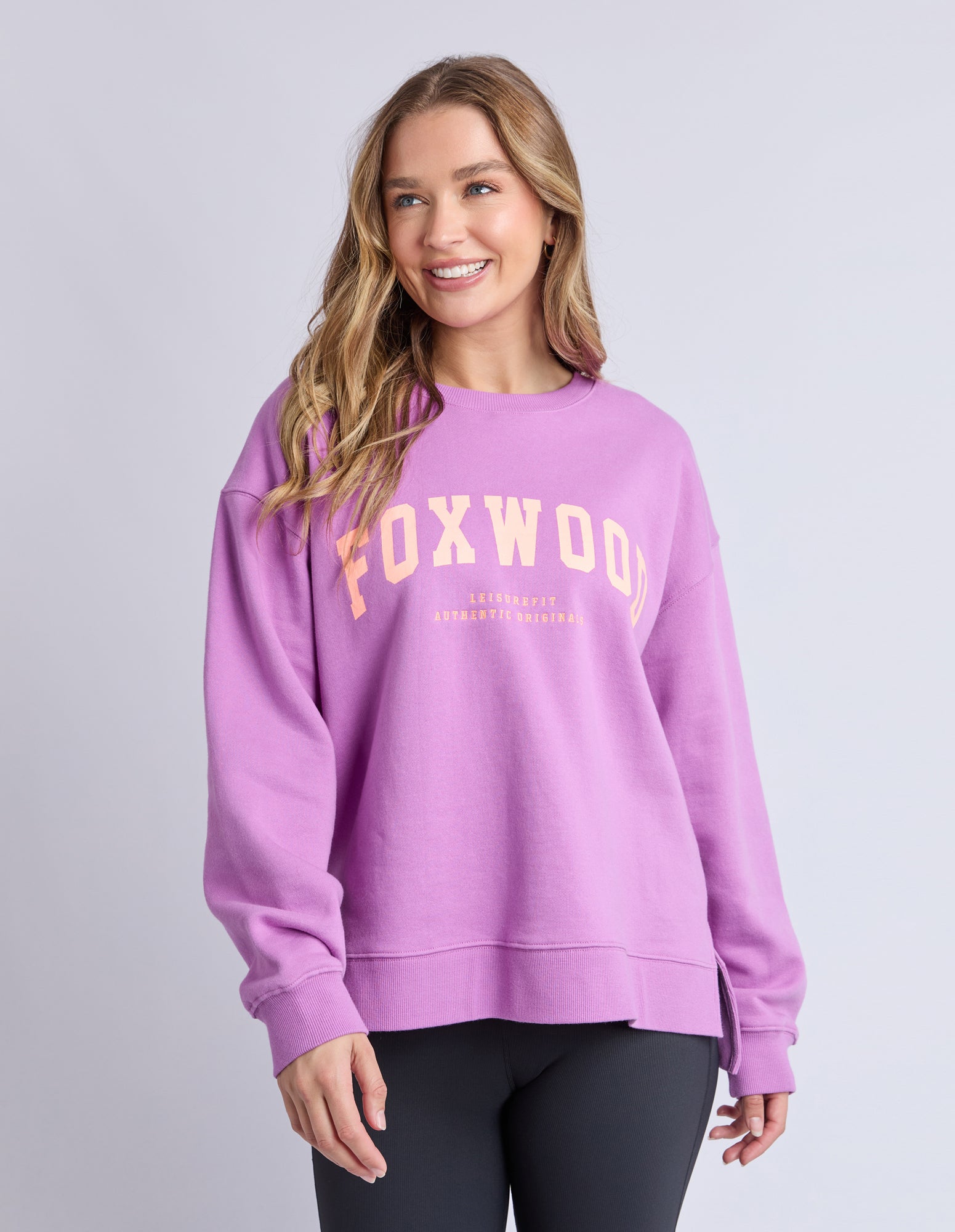 Image of Woman wearing a purple jumper with 'FOXWOOD' printed on it in orange against a grey background