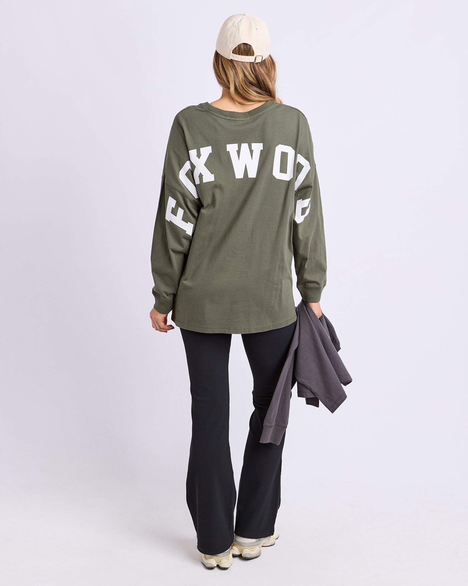 Back view of a woman wearing a khaki  long sleeve tee with 'FOXWOOD' text, black pants, and a white cap on a light grey background