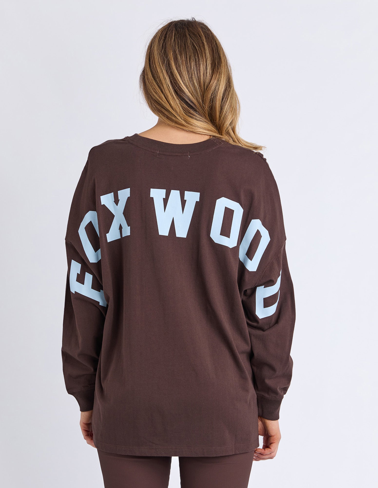 Image of Back view of a woman wearing a brown long sleeve tee with 'FOXWOOD' printed on the back in light blue against a white background 