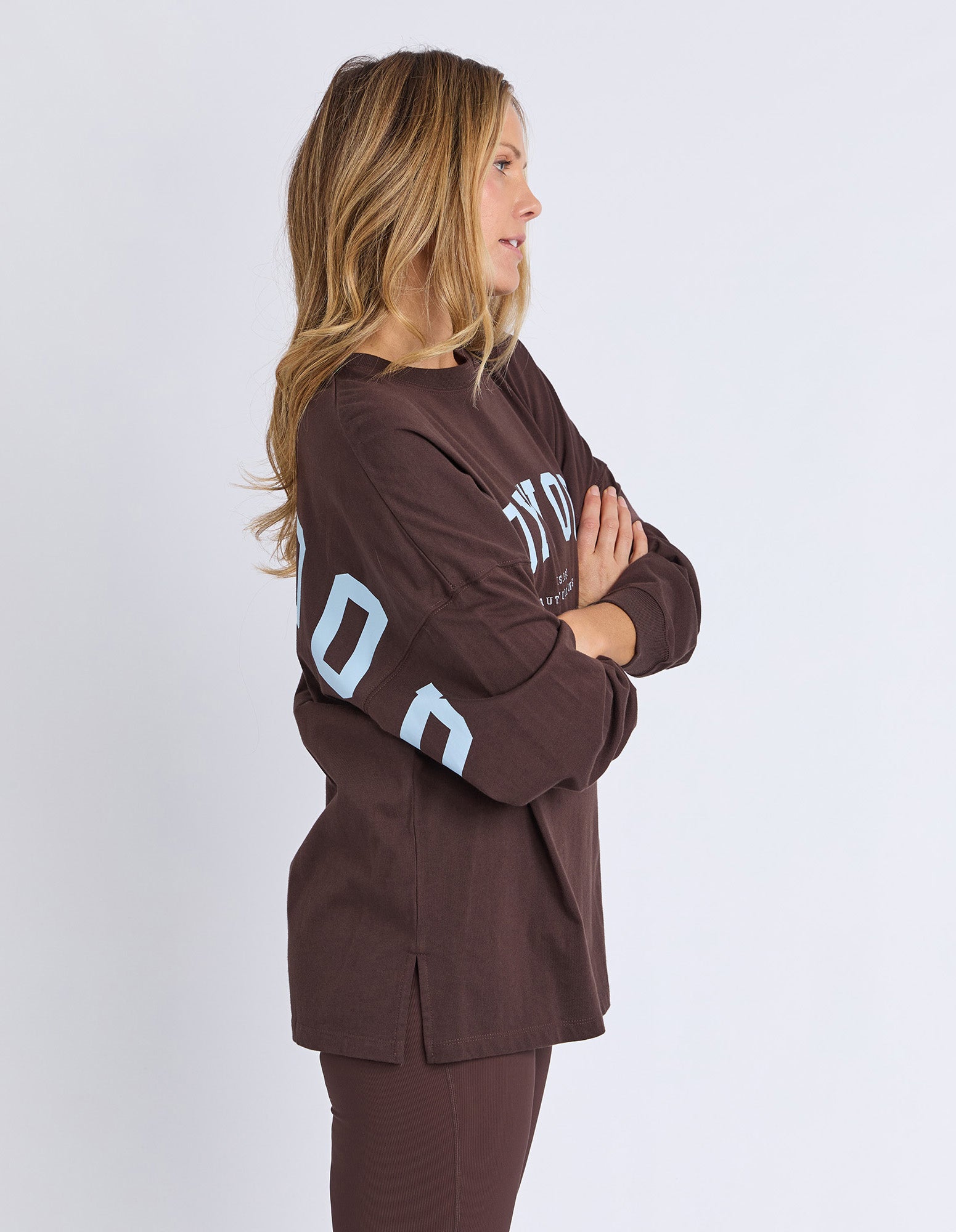 Side view of a woman wearing a brown Foxwood long sleeve tee with text on sleeves in light blue against a white background