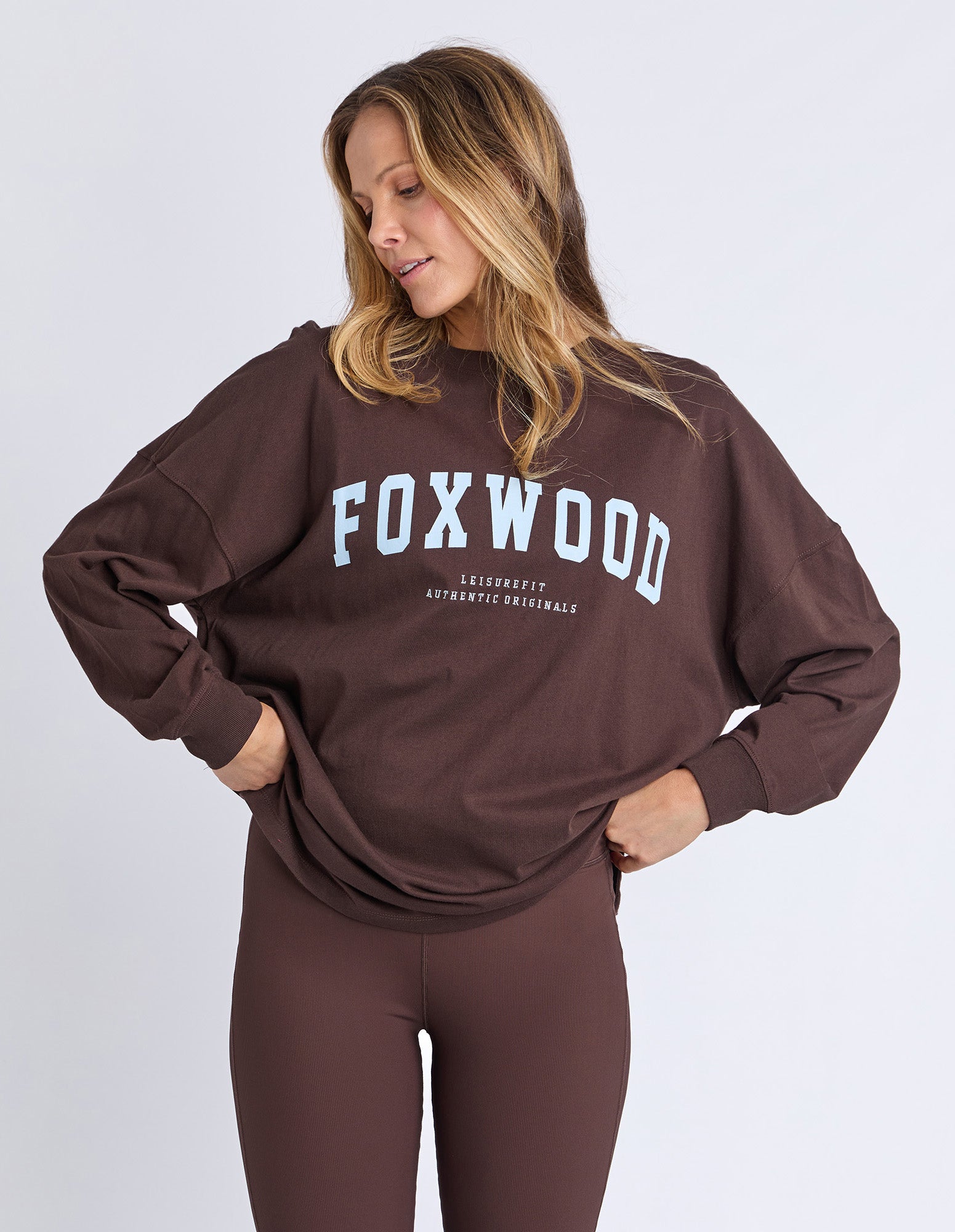 Woman wearing a brown 'FOXWOOD' long sleeve tee with light blue Foxwood logo and leggings on a light grey background