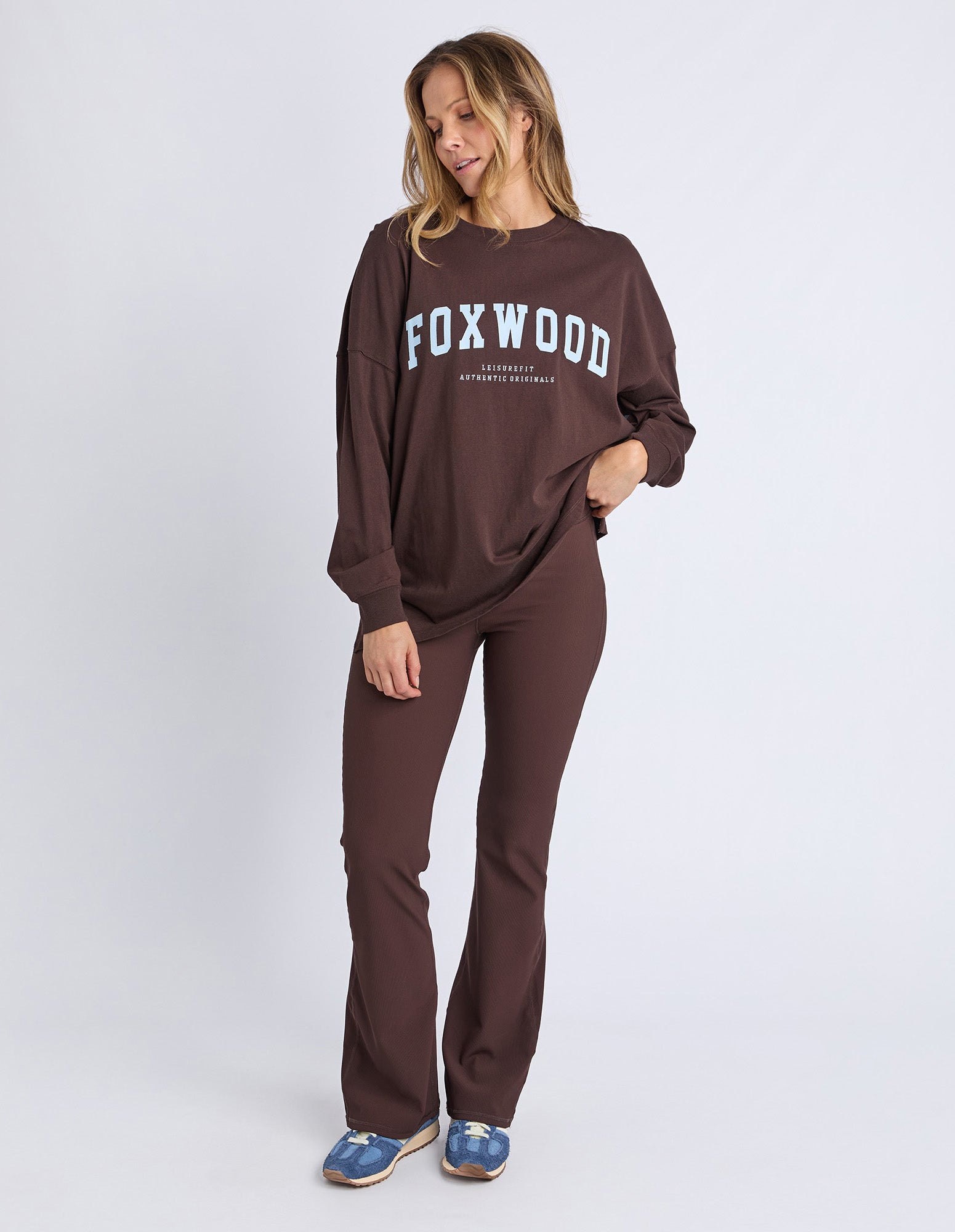 Image of Woman wearing a brown 'FOXWOOD' long sleeve tee with light blue Foxwood logo and pants on a white background