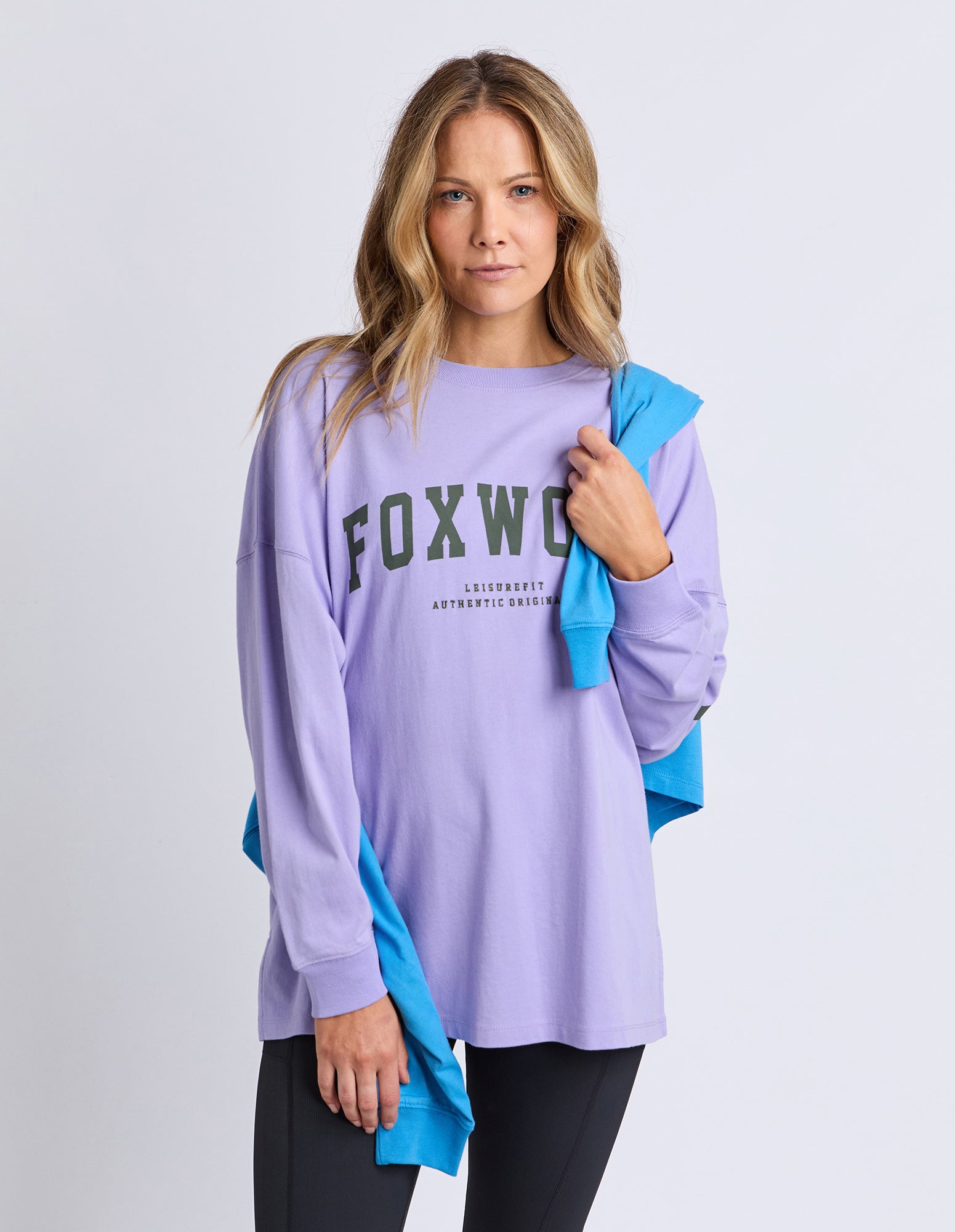 Woman wearing a light purple long-sleeve tee with 'FOXWOOD' branding in khaki with a blue jumper draped over her on a white background