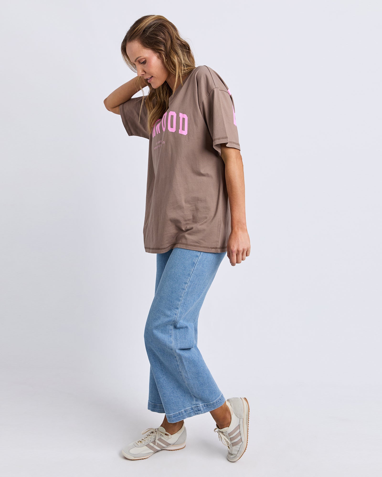 Woman wearing a brown Foxwood oversized t-shirt with 'Foxwood' printed in pink and blue jeans on a white background