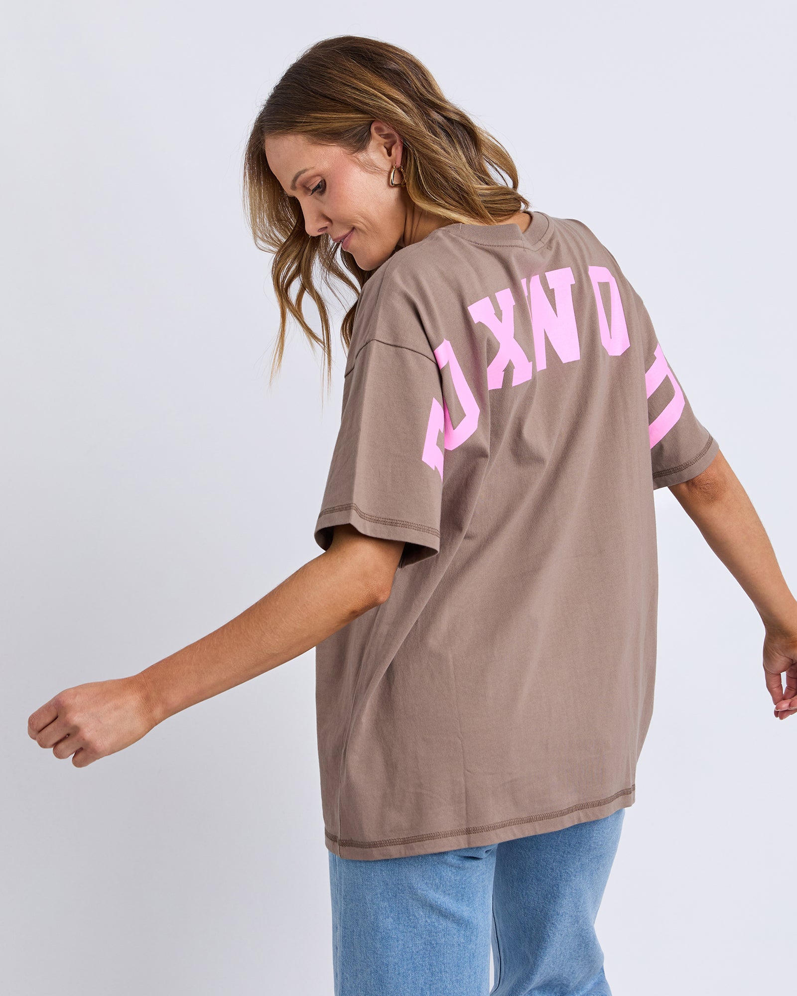 Image of Woman wearing a Foxwood brown oversized t-shirt with pink text on the back against a light grey background