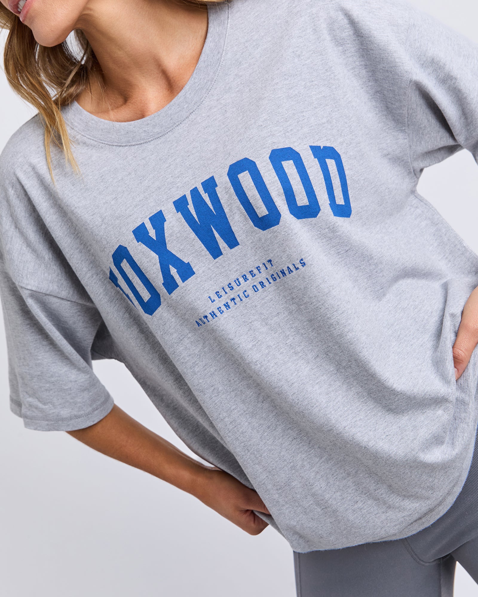 Image of Woman wearing a Grey T-shirt with blue 'FOXWOOD' text on a plain background