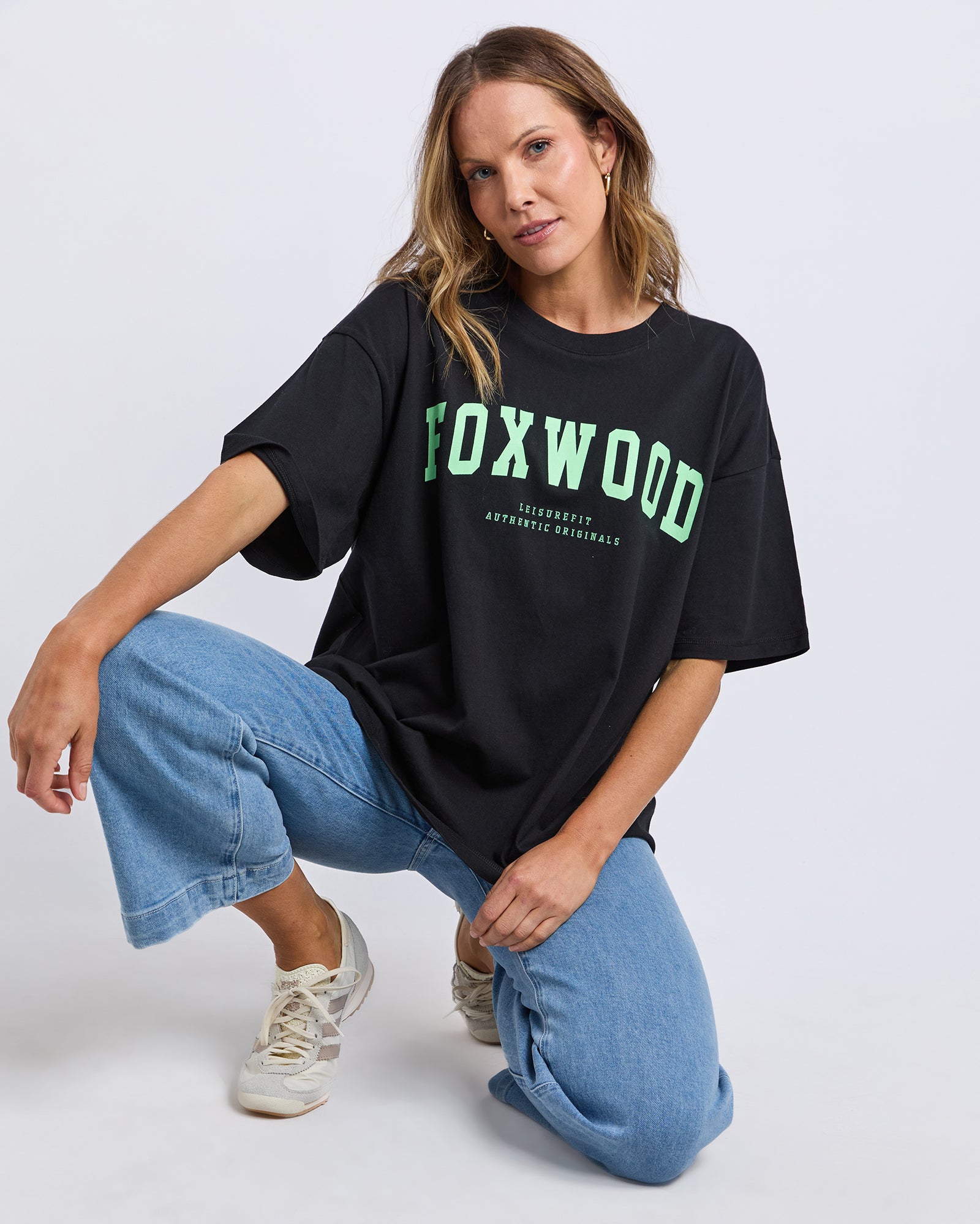 Person wearing a Foxwood black t-shirt with 'FOXWOOD' printed on it in green, sitting on the floor against a white background.