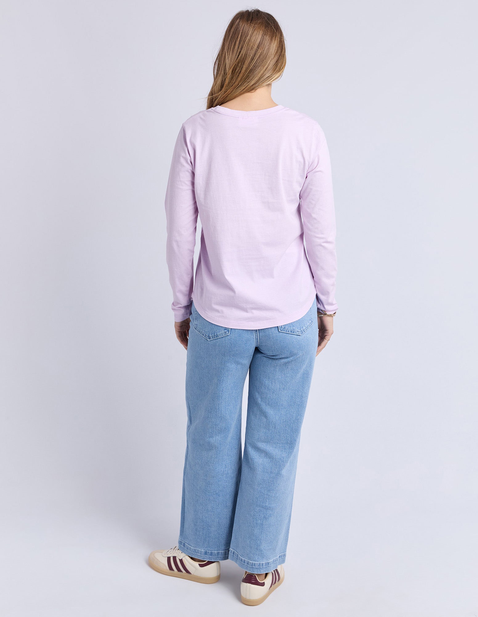 Image of Backview of a woman wearing a light purple long-sleeve tee and blue jeans on a plain background