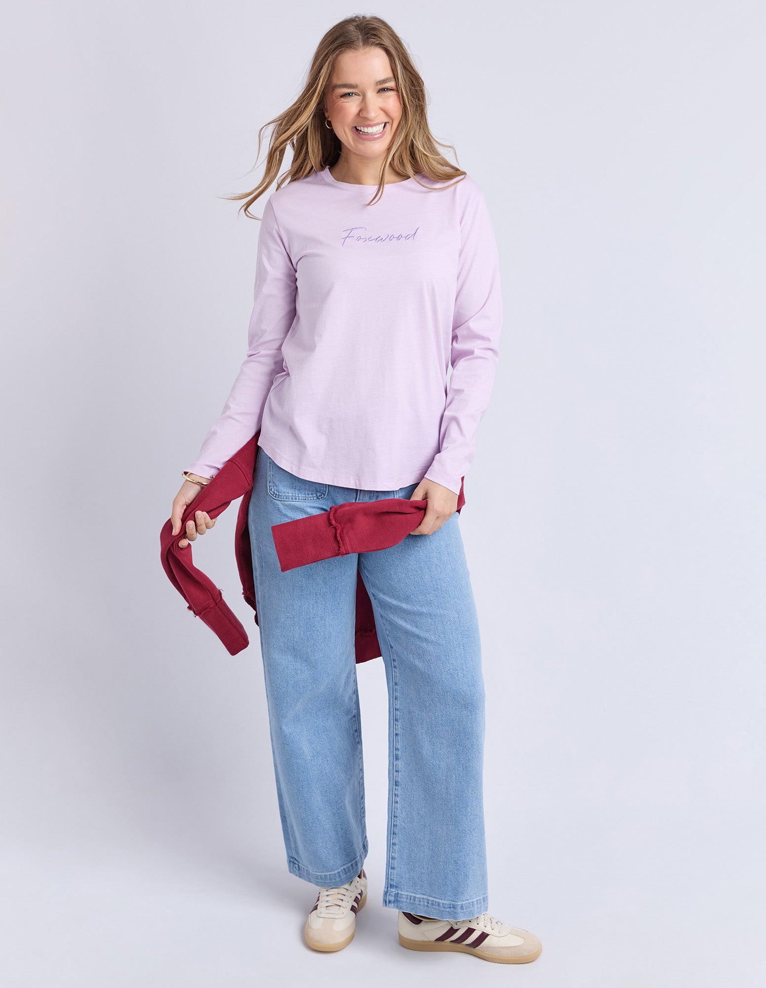 Image of Woman wearing a Foxwood light purple long-sleeve tee and blue jeans, holding a red jacket against a white background.