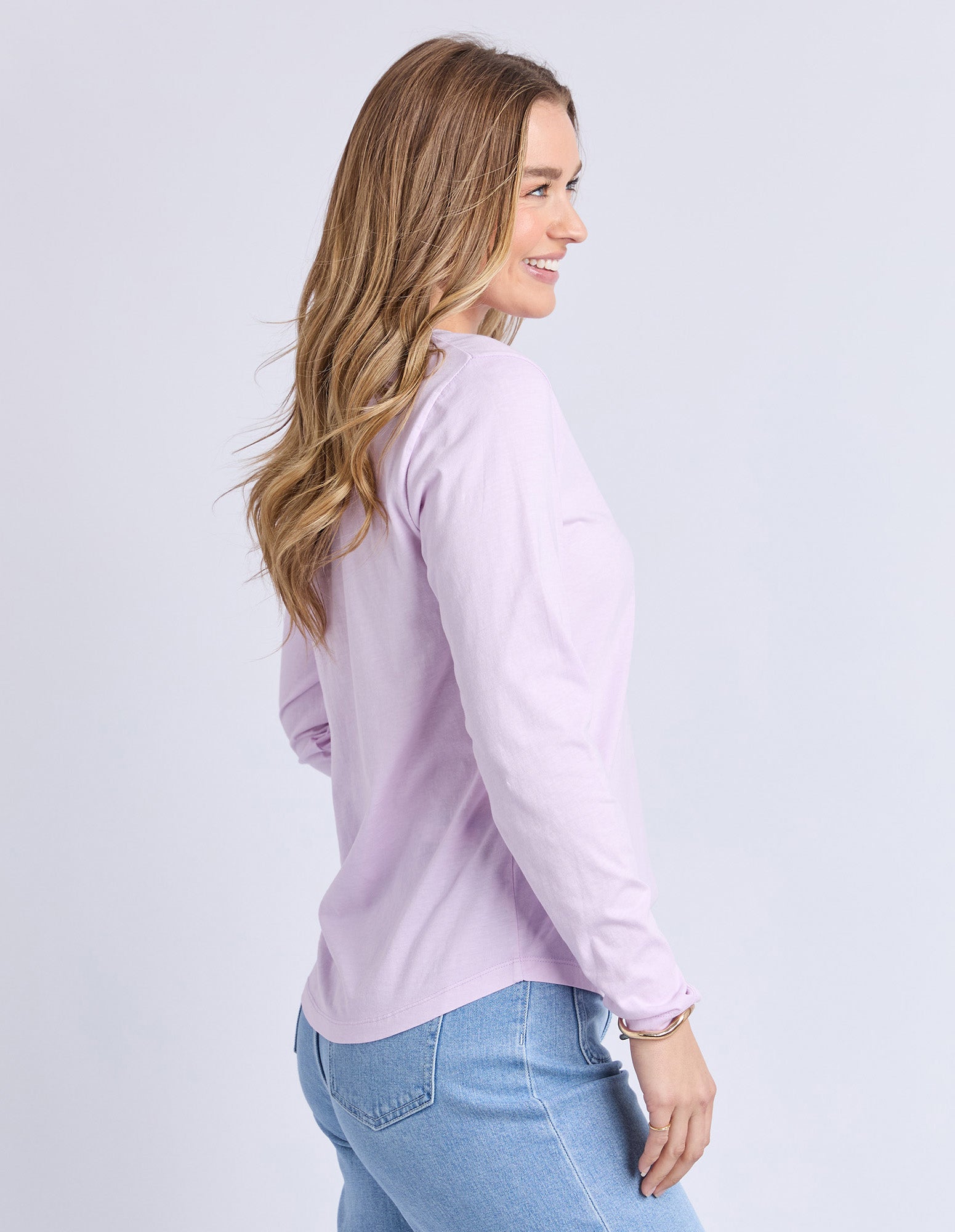 Image of Sideview of a woman wearing a Foxwood light purple long-sleeve tee and blue jeans on a white background
