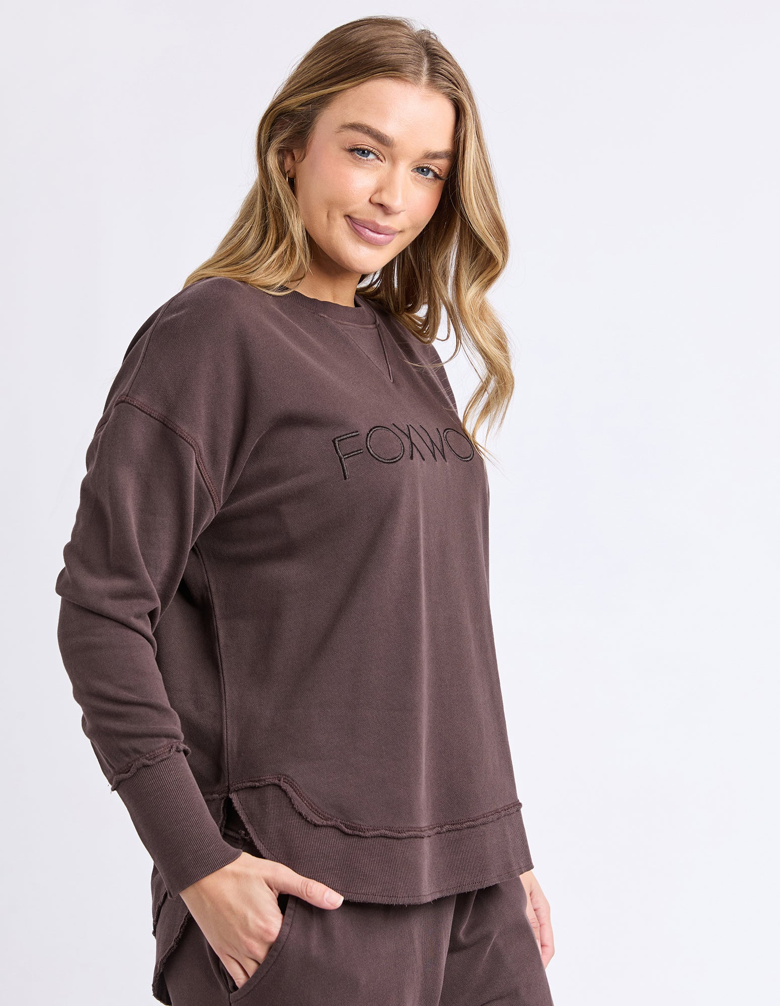Side view of a Woman wearing a brown jumper with 'FOXWOOD' logo on a white background