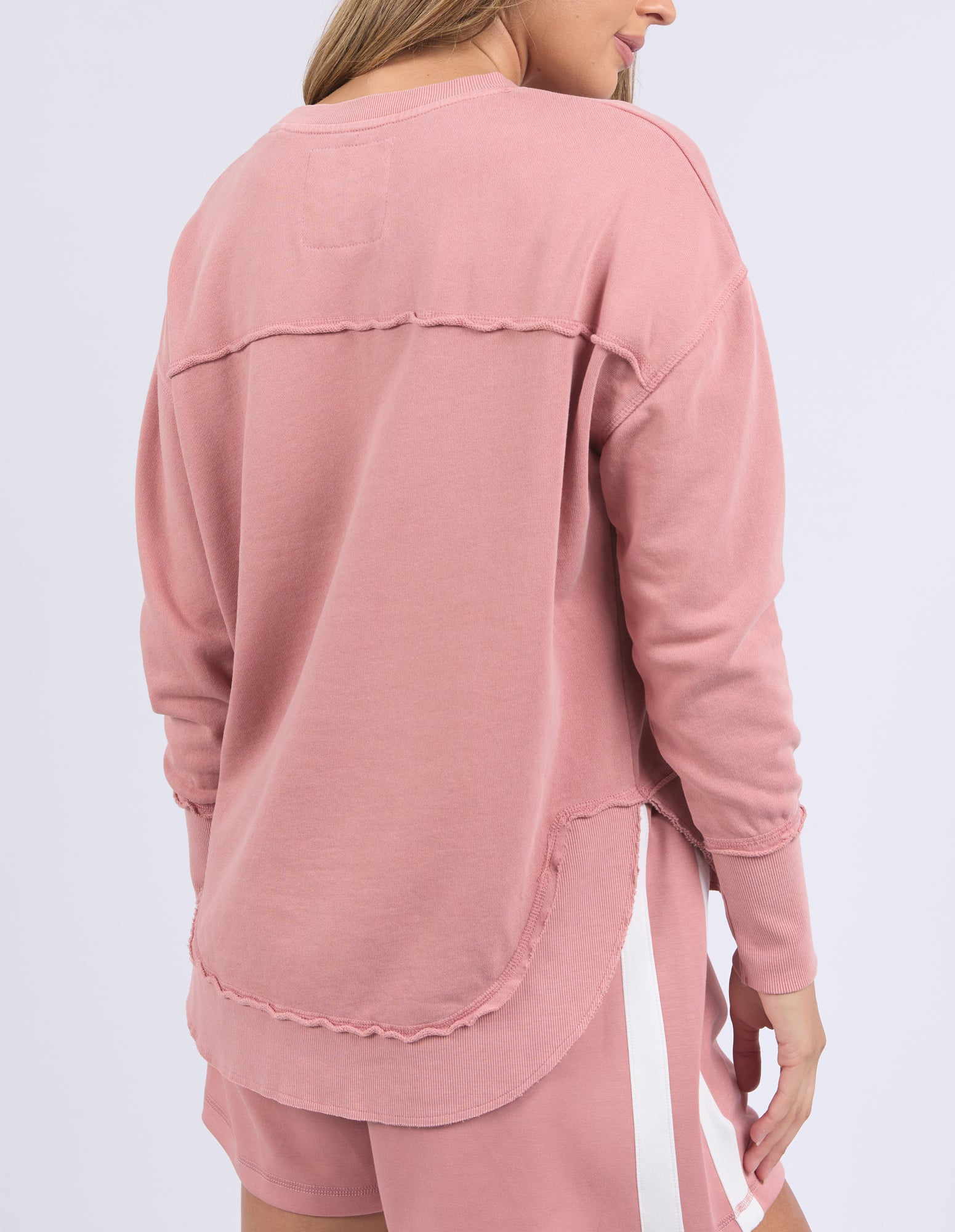 Back view of a woman wearing a dusty rose long-sleeve jumper and shorts on a white background