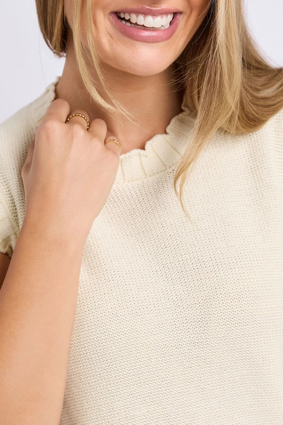 Image of Close-up of a smiling woman wearing a Foxwood cream-colored knit top with a ruffled neckline and short sleeves, accessorized with gold rings.