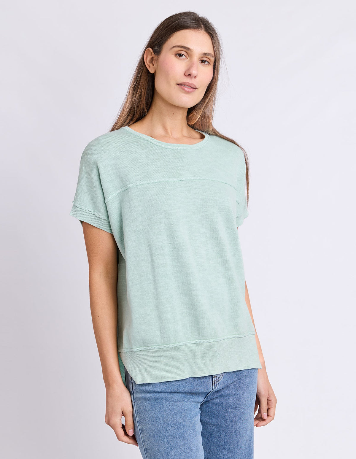 A woman wearing a Foxwood light mint green short-sleeve crewneck top and blue jeans, standing against a plain light gray studio background.