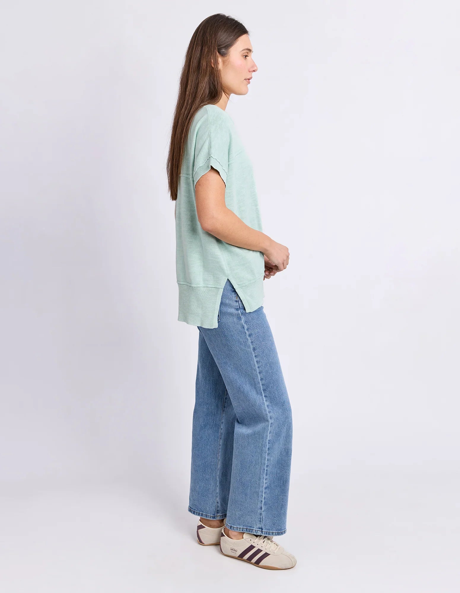 Image of A woman in profile wearing a Foxwood light mint green short-sleeve top, wide-leg blue jeans, and beige sneakers with dark stripes, standing against a plain light gray studio background.