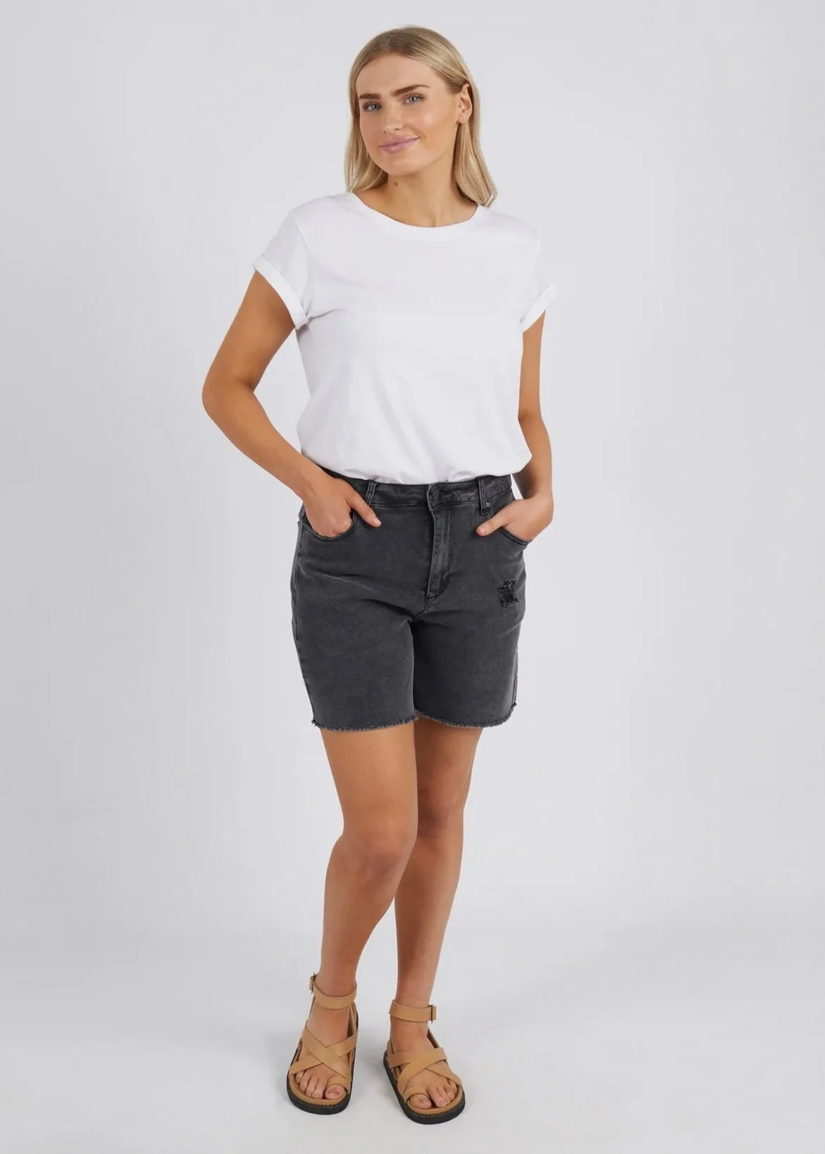 A woman modeling a white short-sleeve crewneck t-shirt and Foxwood dark gray denim shorts, paired with tan leather sandals, against a light gray studio background.