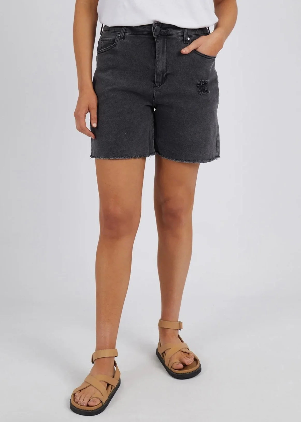Image of A woman wearing Foxwood dark gray washed denim shorts with a frayed hem, paired with tan slide sandals and a white top, shown from the waist down against a light gray studio background.