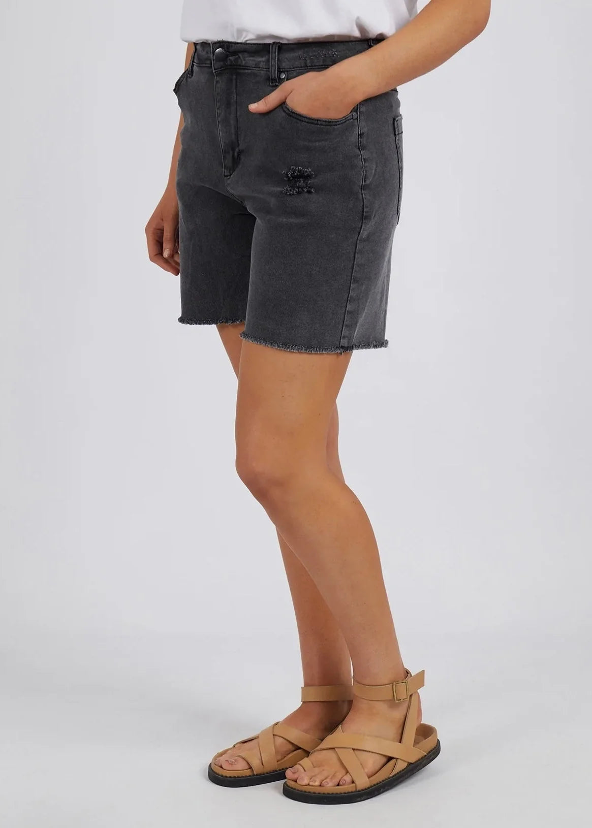 Image of A woman wearing Foxwood dark gray denim shorts with a distressed logo patch on the left thigh, paired with tan leather sandals and a white top, shown from the waist down against a light gray studio background.