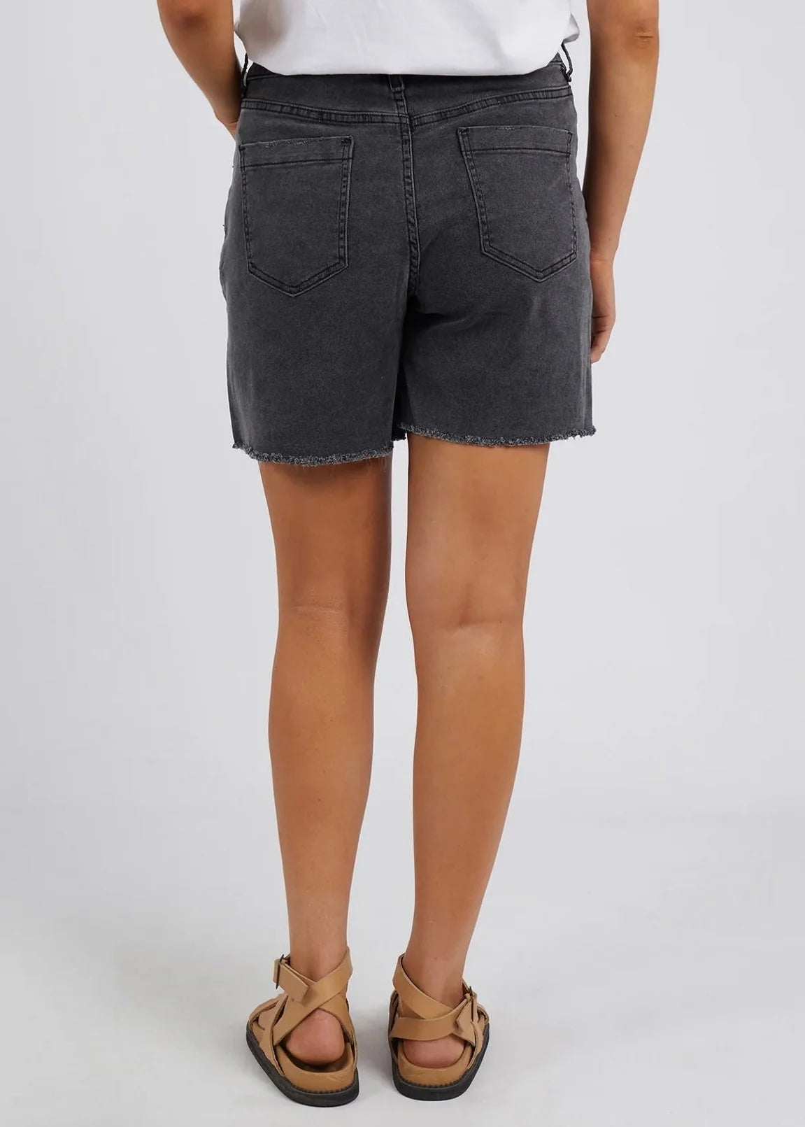 Image of Rear view of a woman wearing Foxwood dark gray washed denim shorts with a frayed hem, paired with a white top and tan sandals, against a light gray studio background.