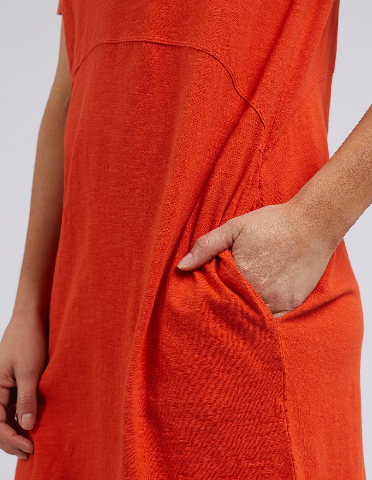 Close-up of a woman wearing a bright Foxwood orange-red linen dress with a side pocket, showing the fabric texture and a hand in the pocket.