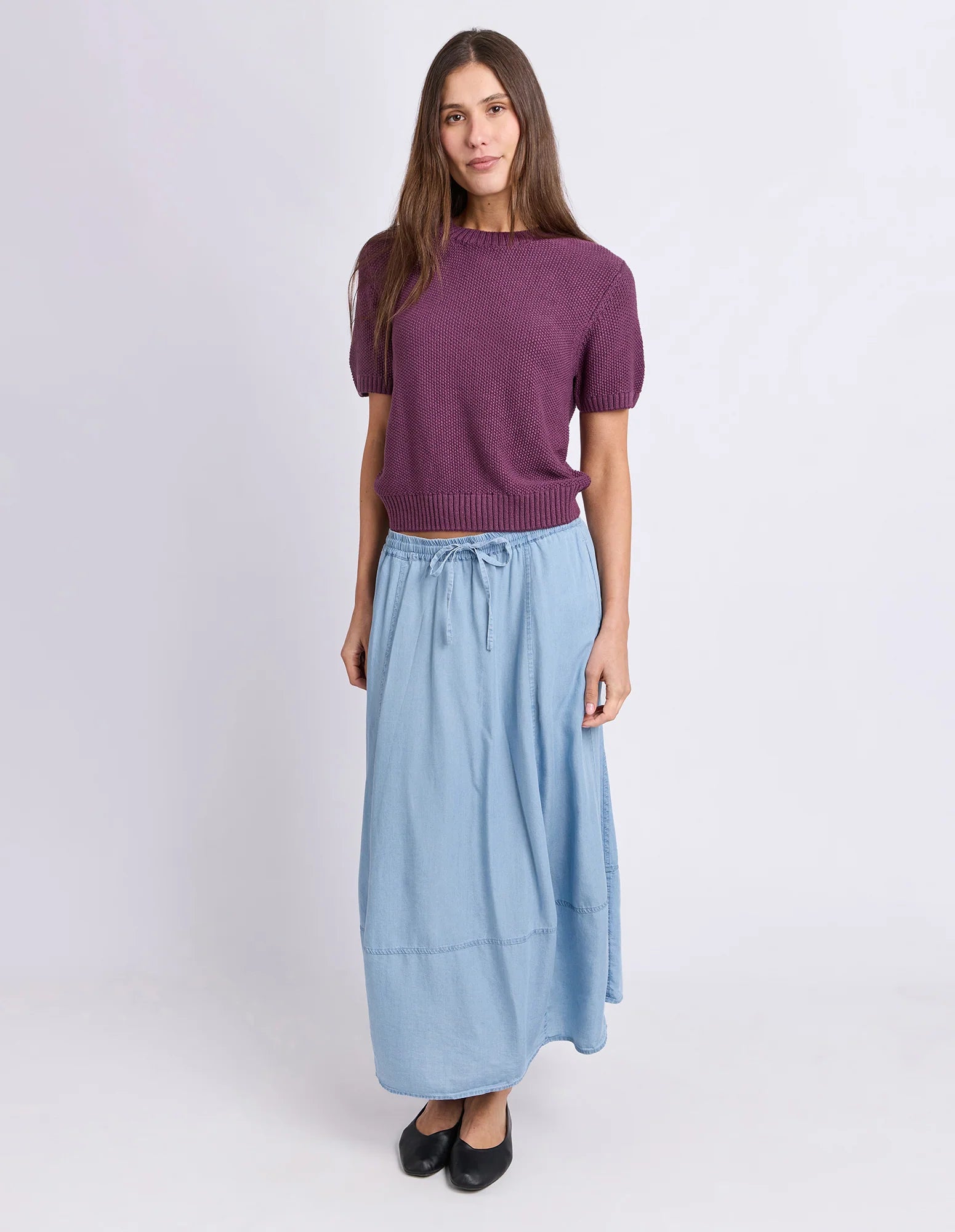 Image of A woman modeling a Foxwood short-sleeve purple knit sweater paired with a light blue midi skirt and black flats, standing against a plain light gray studio background.